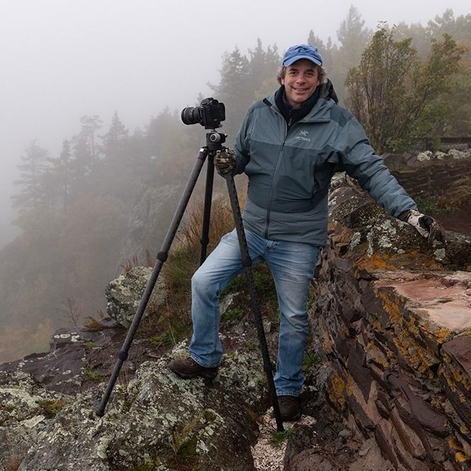 A photographer with camera and tripod stands on a misty mountain ledge during an outdoor photo session.