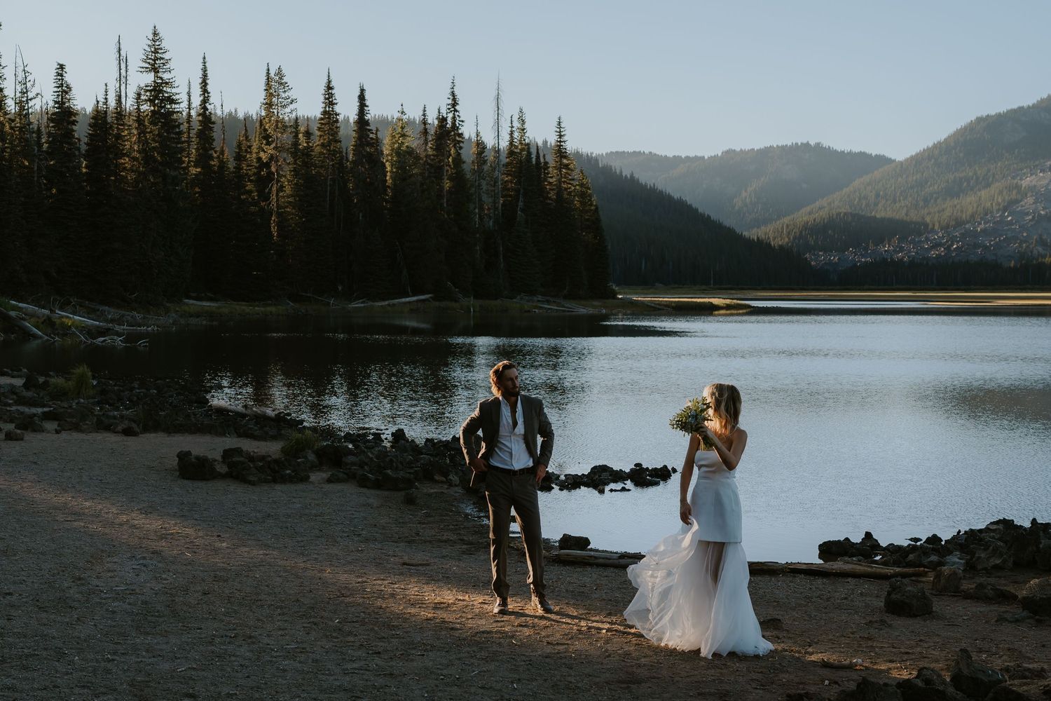 A couple stands at the edge of a serene mountain lake surrounded by evergreen forest at a sunset elopement.