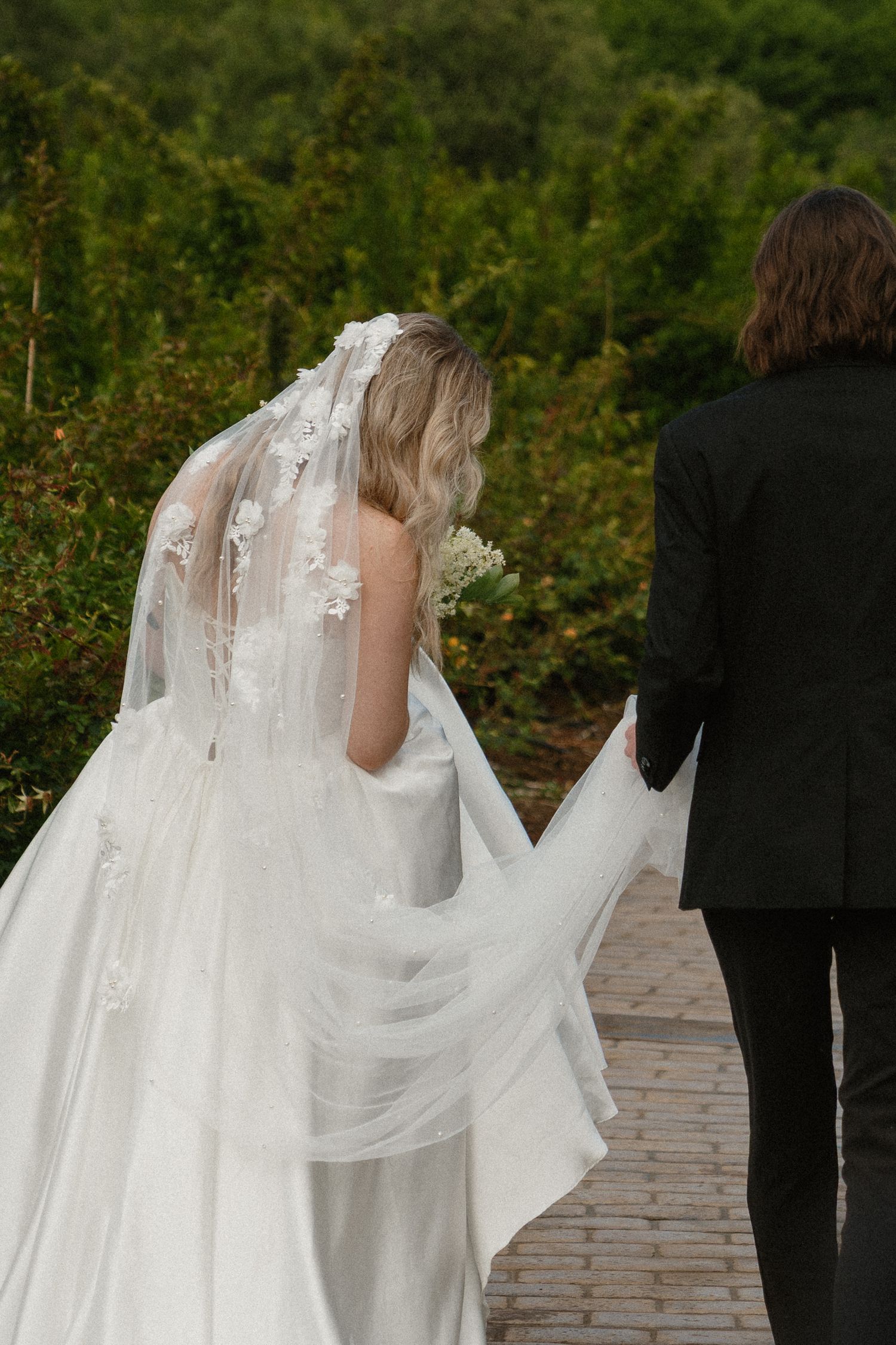 A bride in a flowing white dress with floral veil walks along a brick path with another person in black attire.