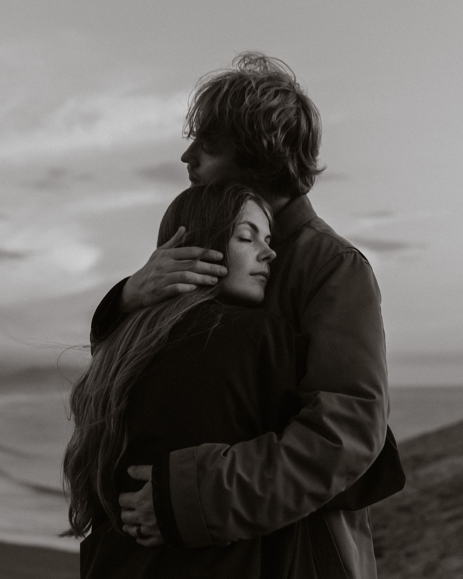 A couple in dark clothing embraces intimately against a moody cloudy sky in black and white.