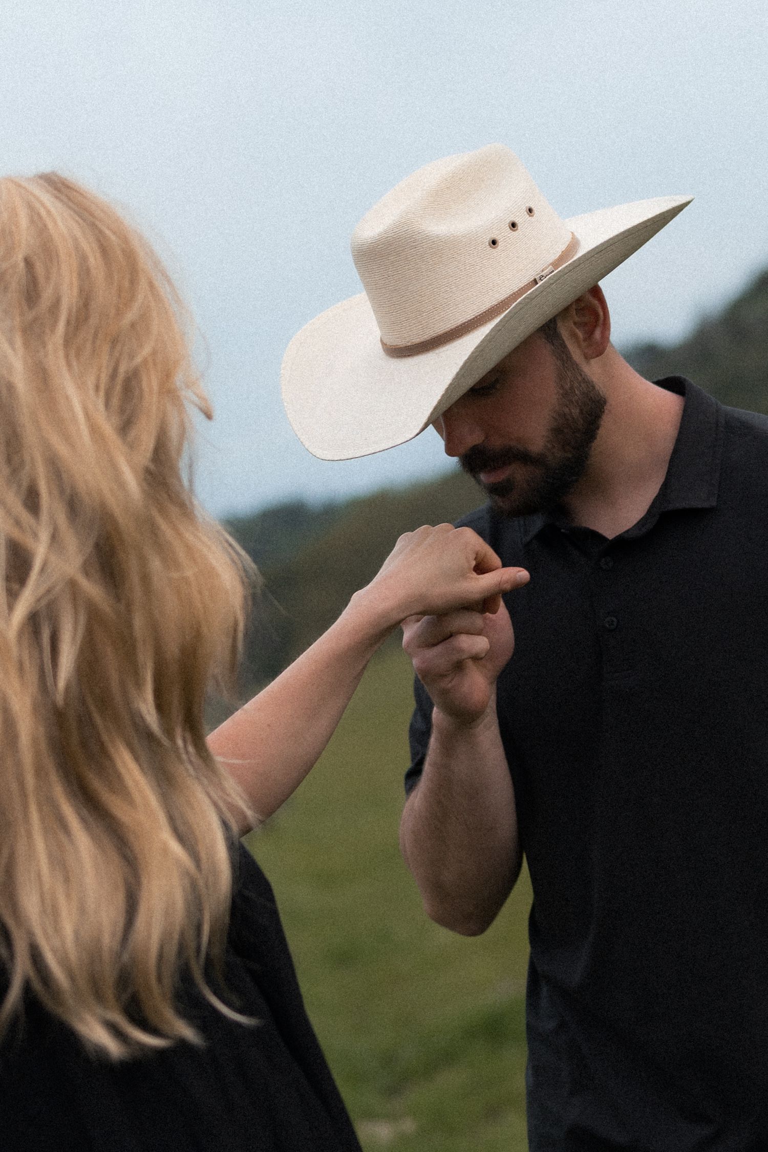 A romantic moment captured outdoors as a person in a cowboy hat tenderly kisses someone's hand in a grassy field.