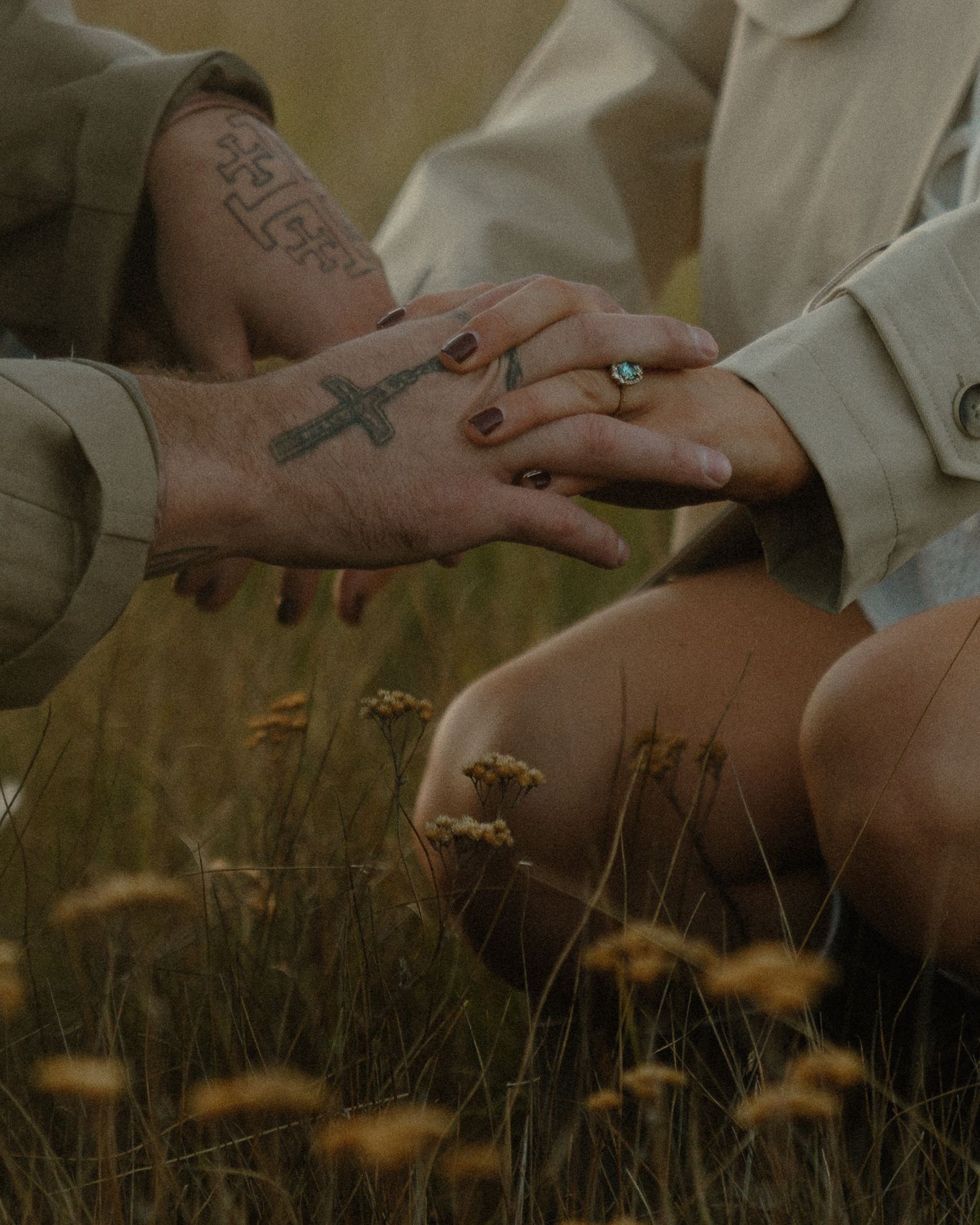Two hands gently touch in a field of tall grass and wildflowers during golden hour.