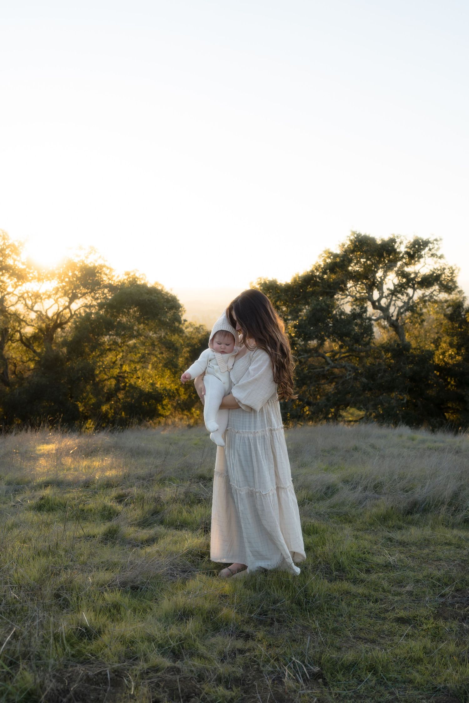 A young mother in long white dress holding an infant in golden evening light amidst tall grass and trees at sunset.