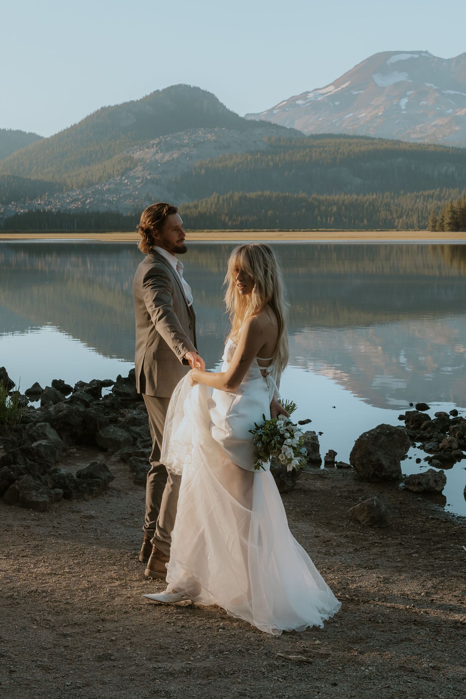 A couple shares an intimate moment by a serene mountain lake at sunset, with the bride wearing a flowing white gown.