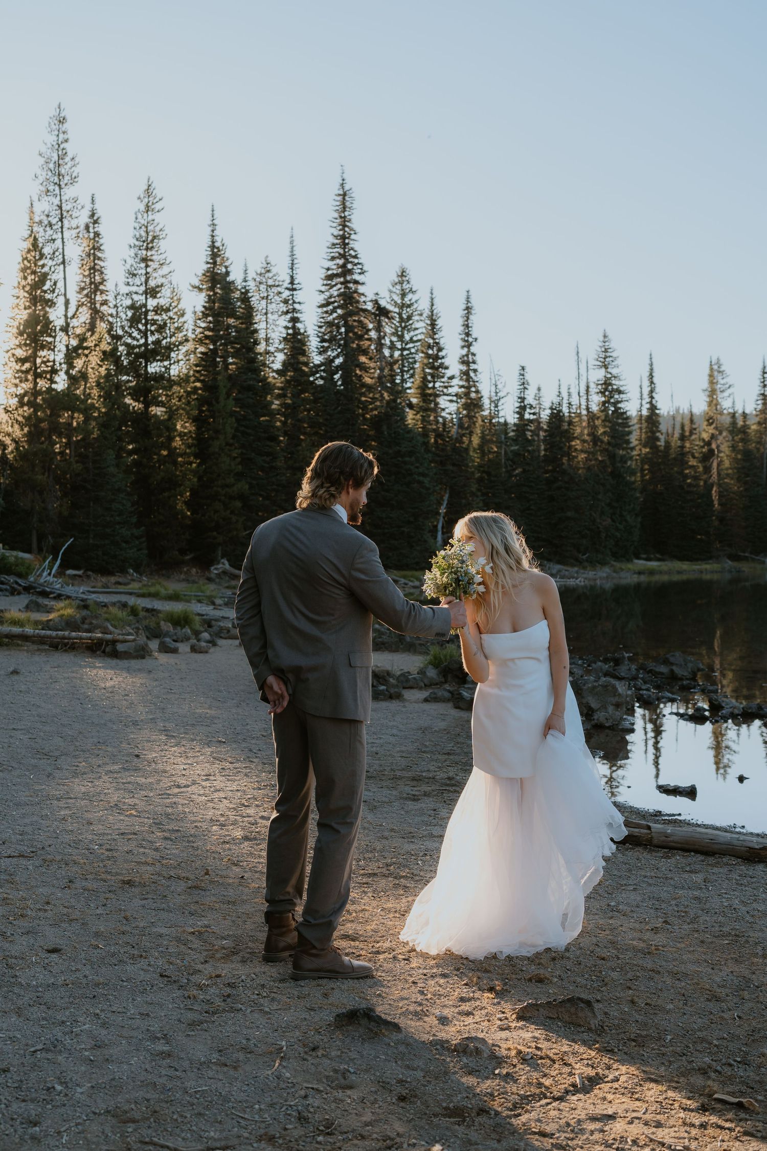 A couple in wedding attire stands by a lake at sunset with evergreen trees silhouetted in the background.