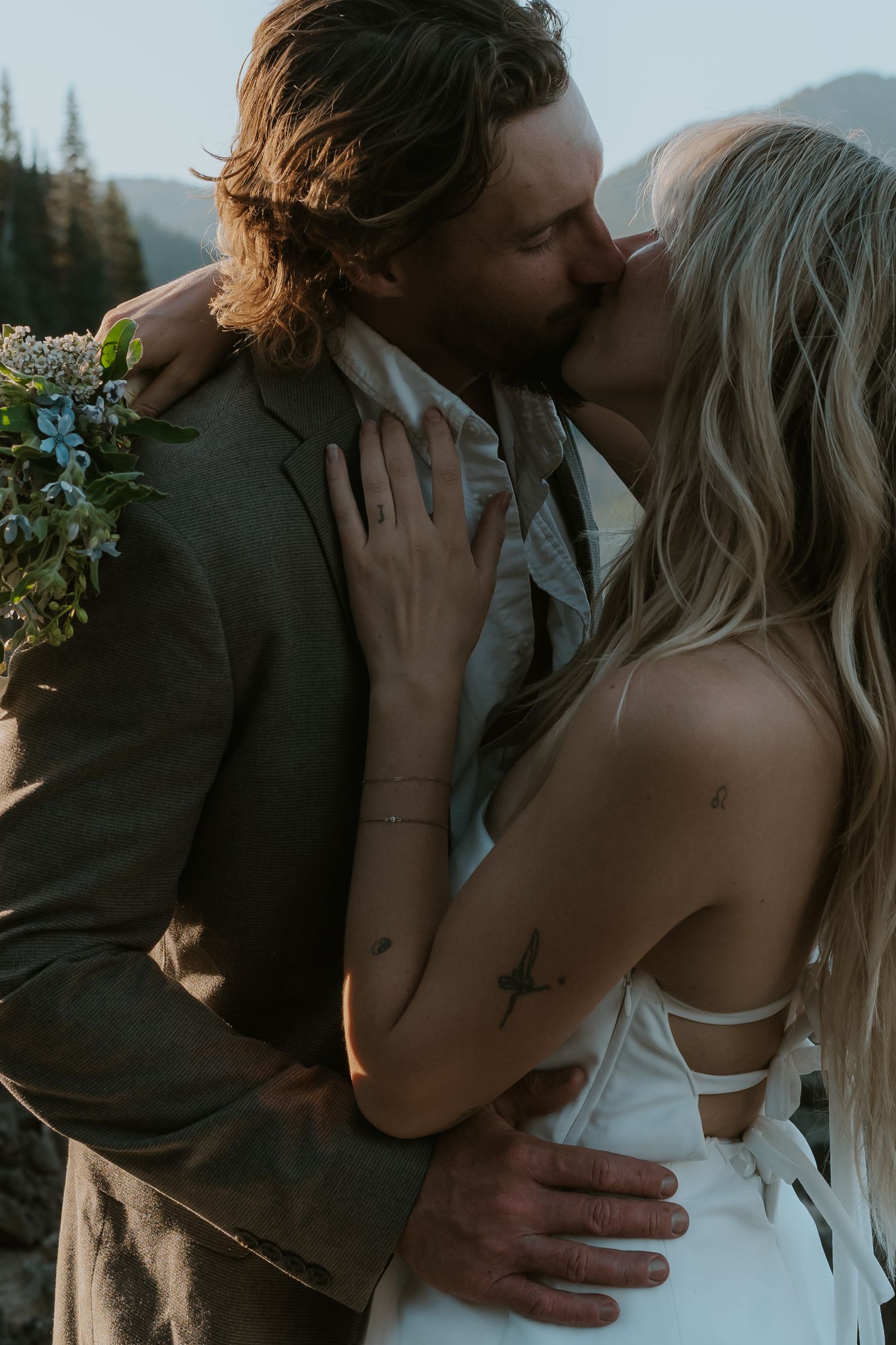 A romantic wedding couple shares an intimate kiss in a mountain setting at sunset with a delicate floral bouquet.