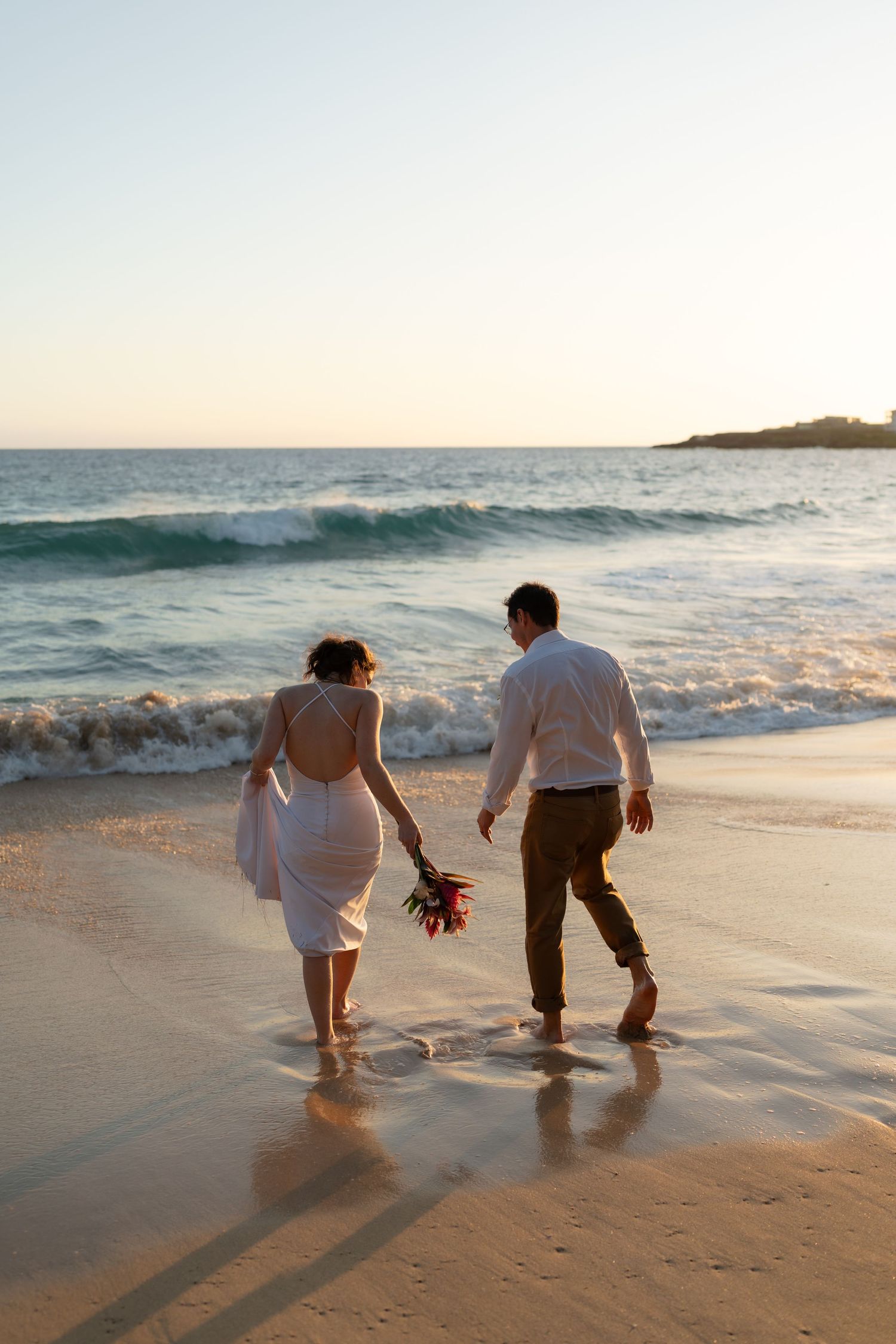 A couple walking together on a sandy beach at their sunset elopement, while waves crash against the shore in the background.