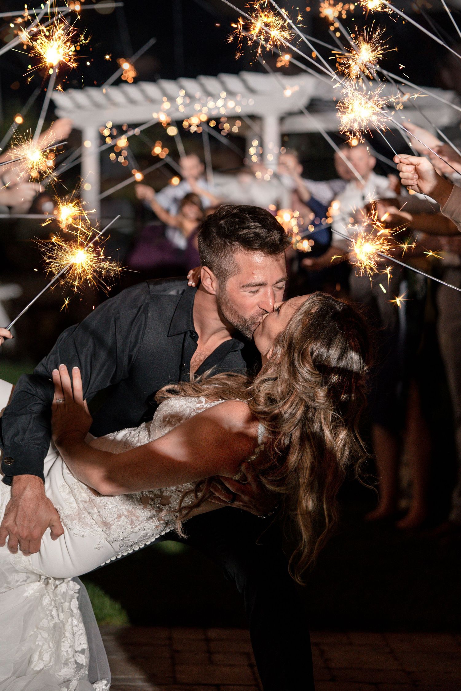 Romantic couple sharing a kiss under sparkling wedding sparklers during an outdoor evening celebration beneath a pergola.