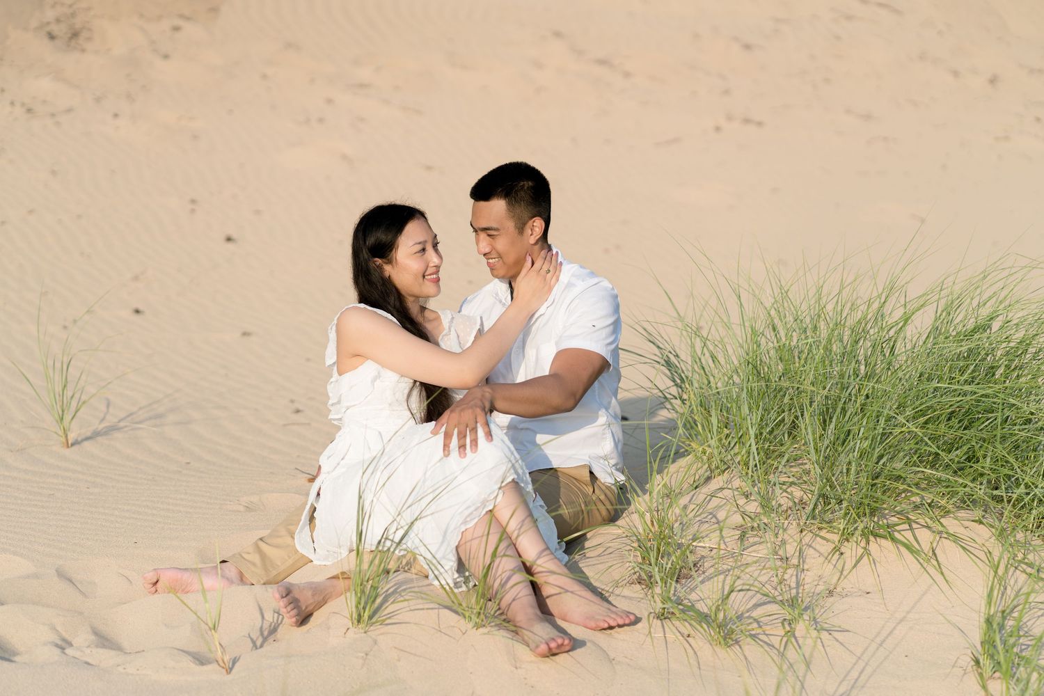 A couple in white outfits sitting together on sand dunes with beach grass in a romantic outdoor setting.