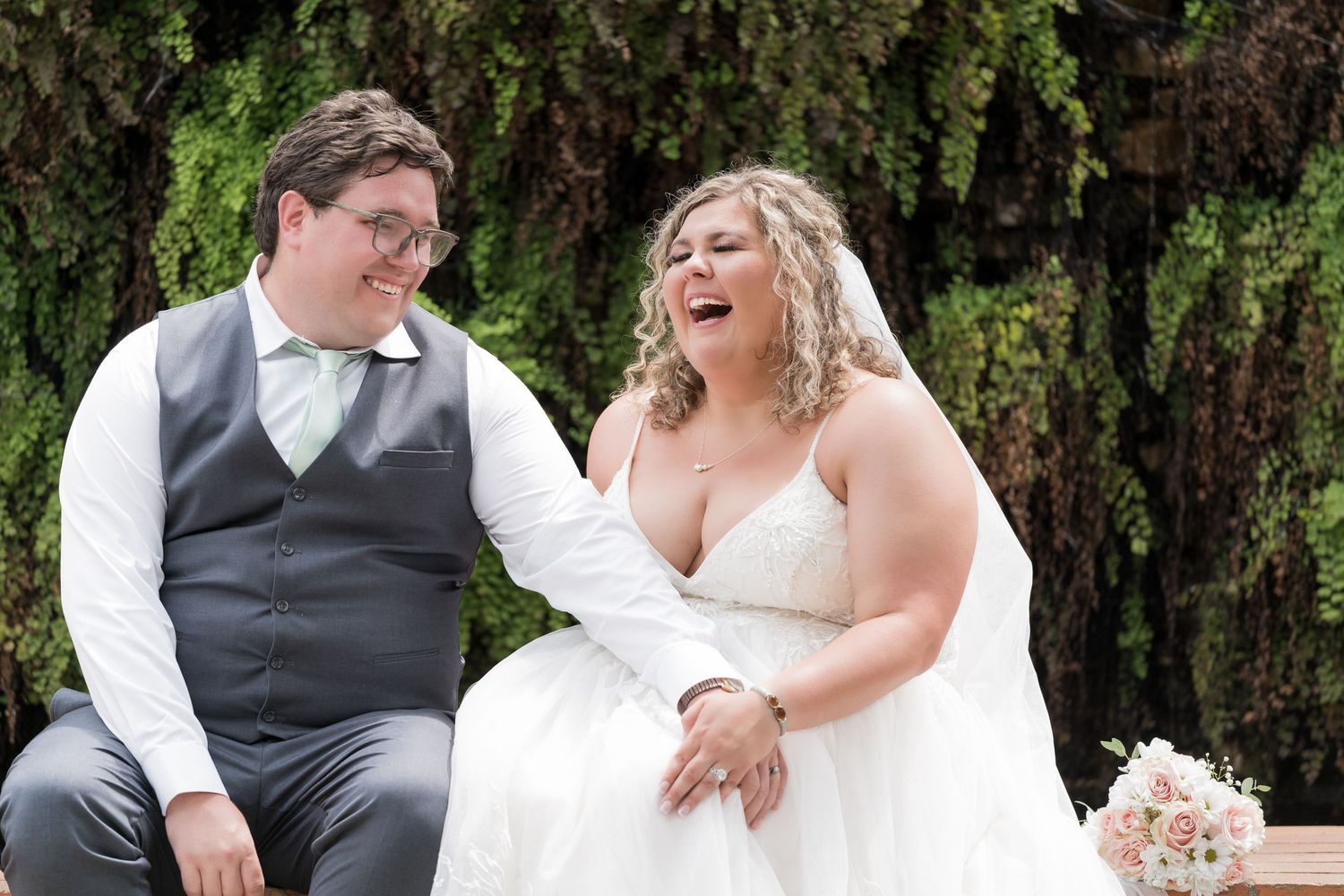 A joyful wedding couple sharing a laugh together while seated outdoors with greenery and flowers in the background.