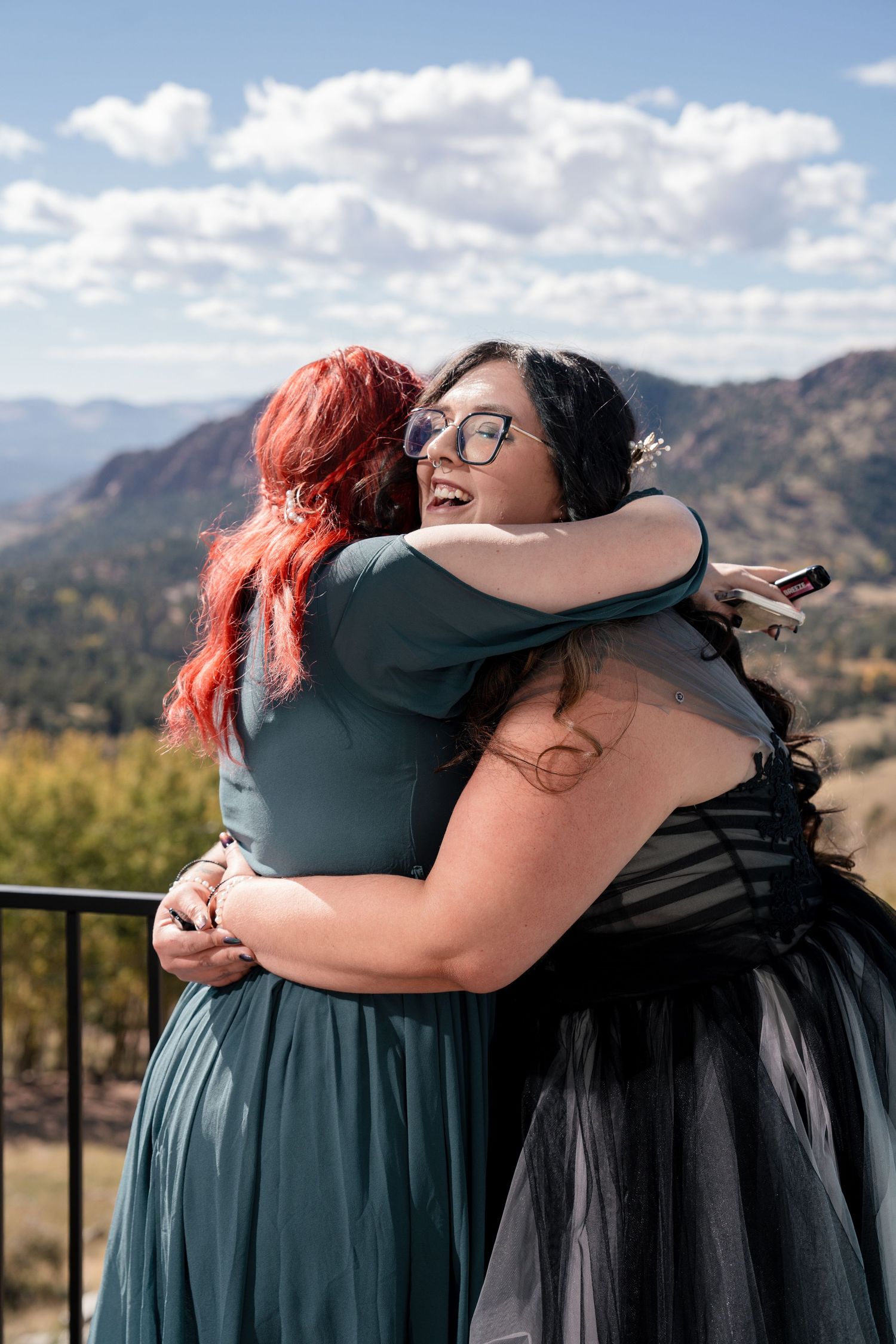 Two friends share a joyful embrace while wearing elegant dresses against a backdrop of mountains and blue sky.
