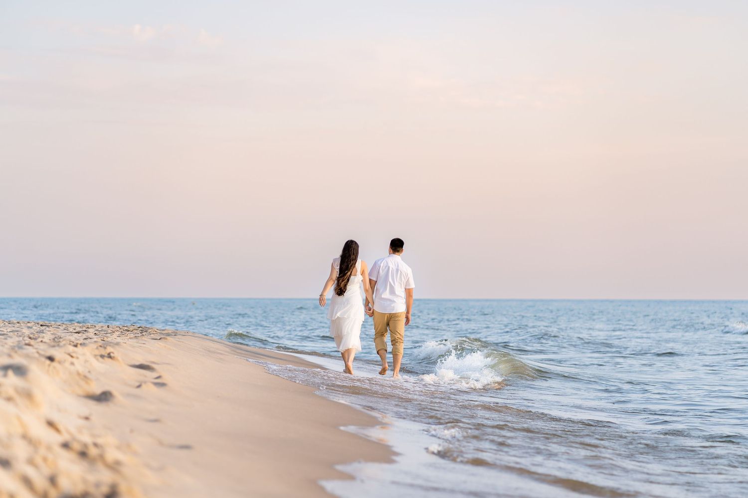 A couple strolls hand in hand along a serene beach at sunset with gentle waves lapping at the shore.