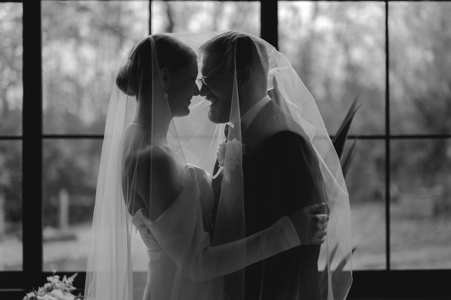 A romantic wedding couple shares a tender moment in silhouette against a window backdrop.
