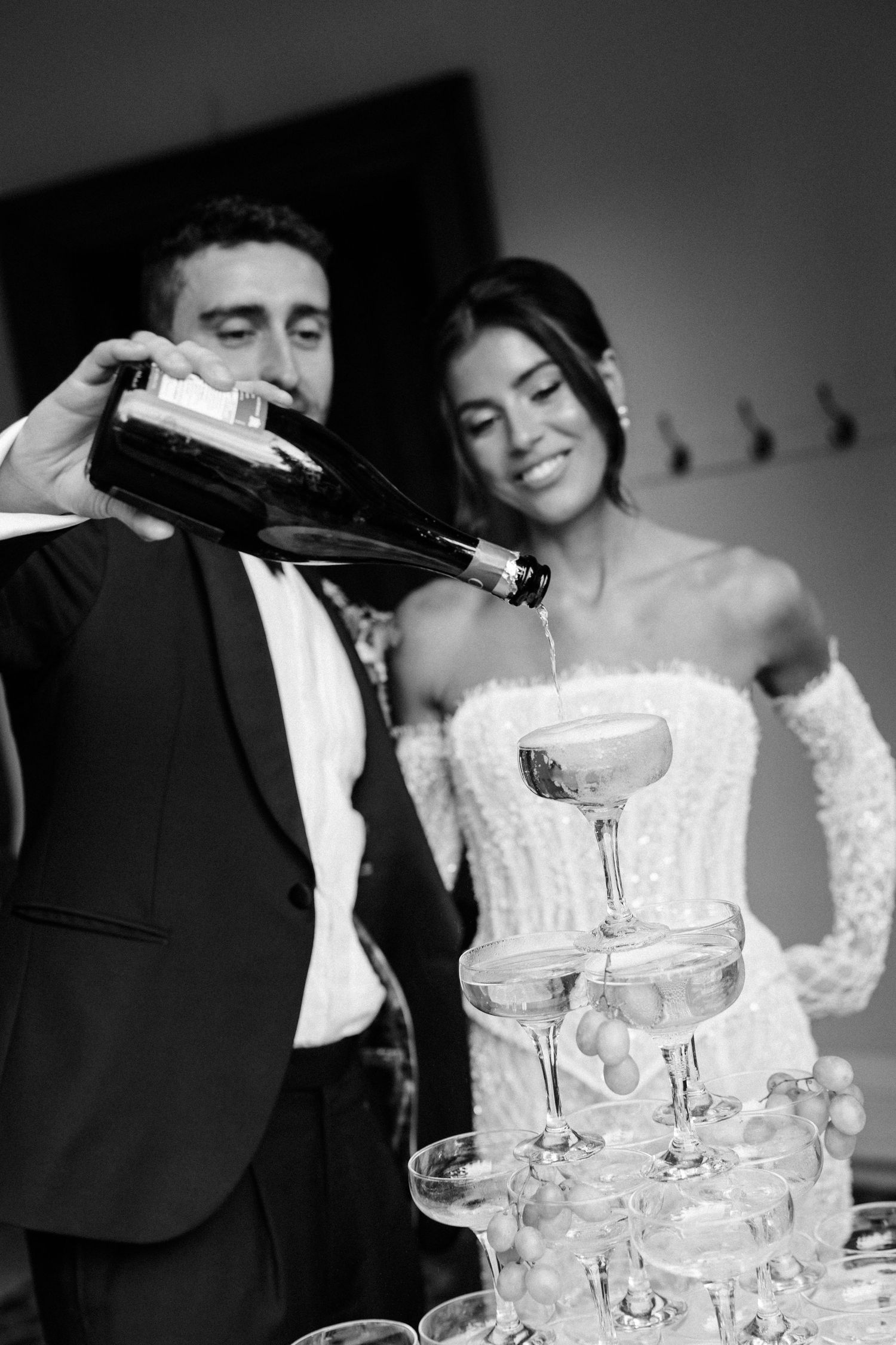 Black and white wedding photos show couple pouring champagne into a tower of glasses at their reception celebration.