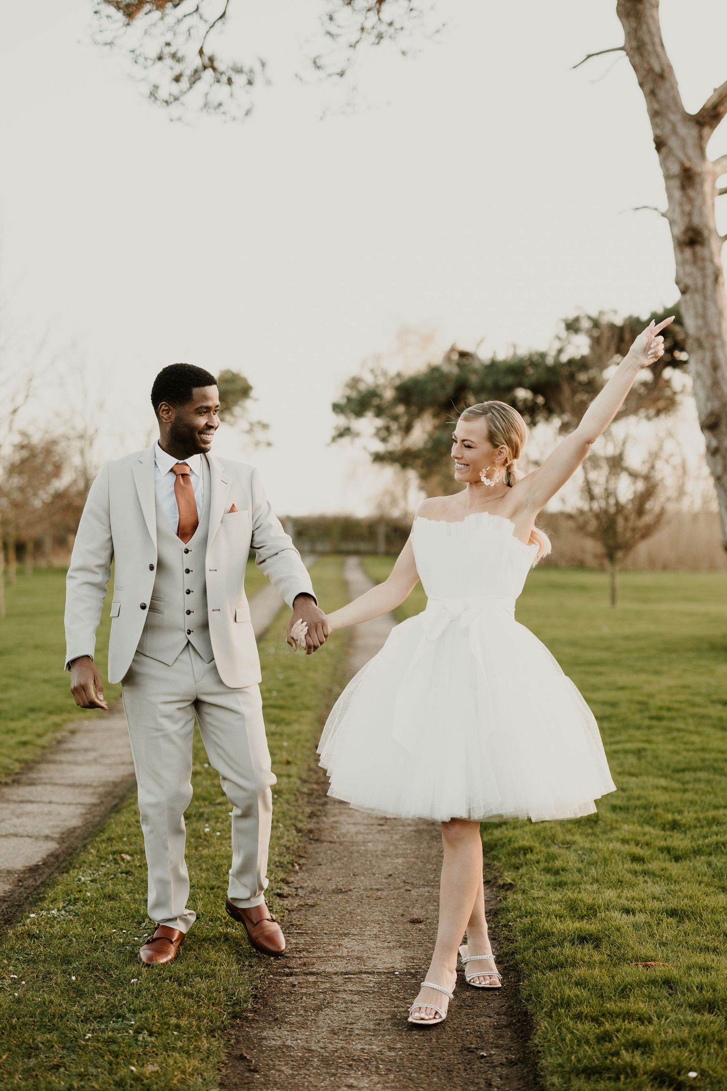 A couple walks and dances playfully along a dirt path through a grassy field at sunset in coordinated wedding attire.
