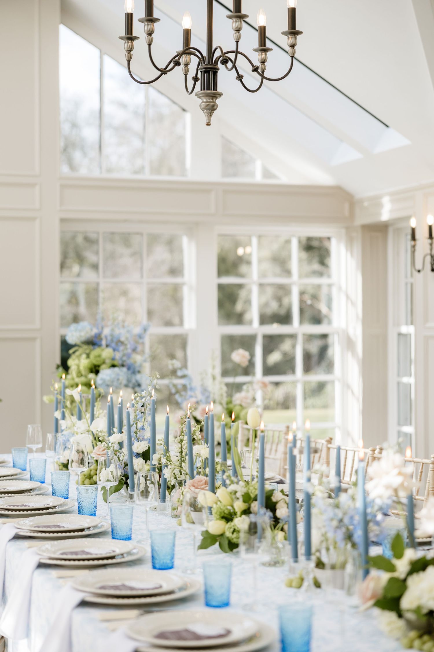Elegant wedding reception table setting with blue glassware, white flowers and chandeliers in bright sunlit room.