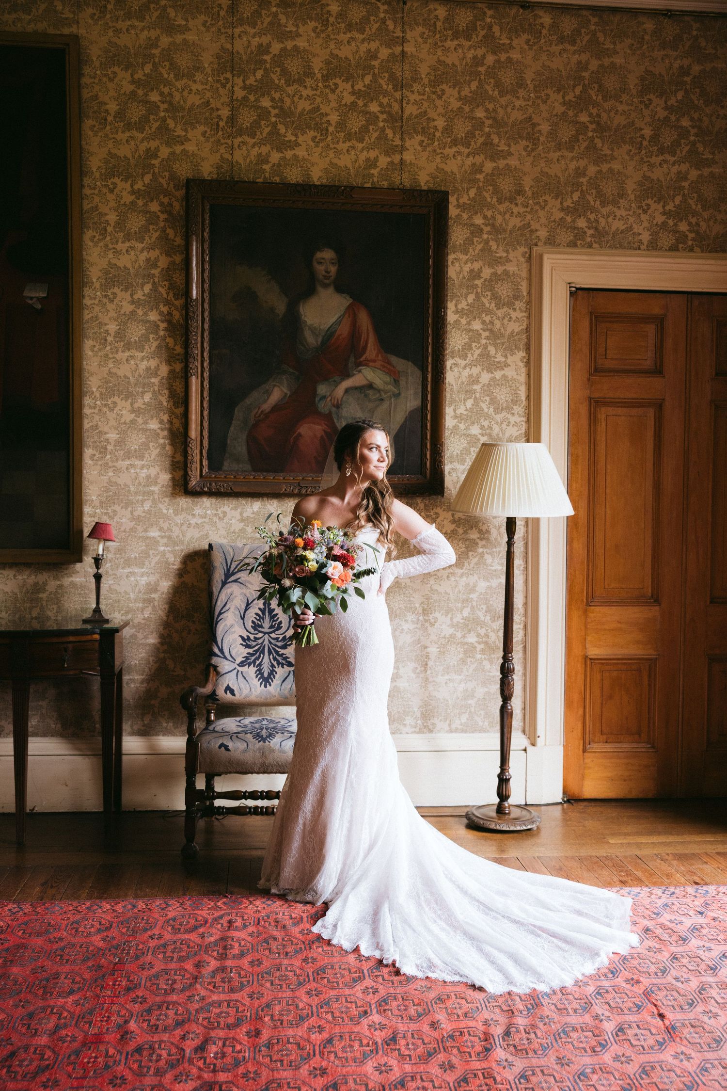 A bride in a fitted lace gown poses elegantly in a stately room with vintage decor and a red carpet.