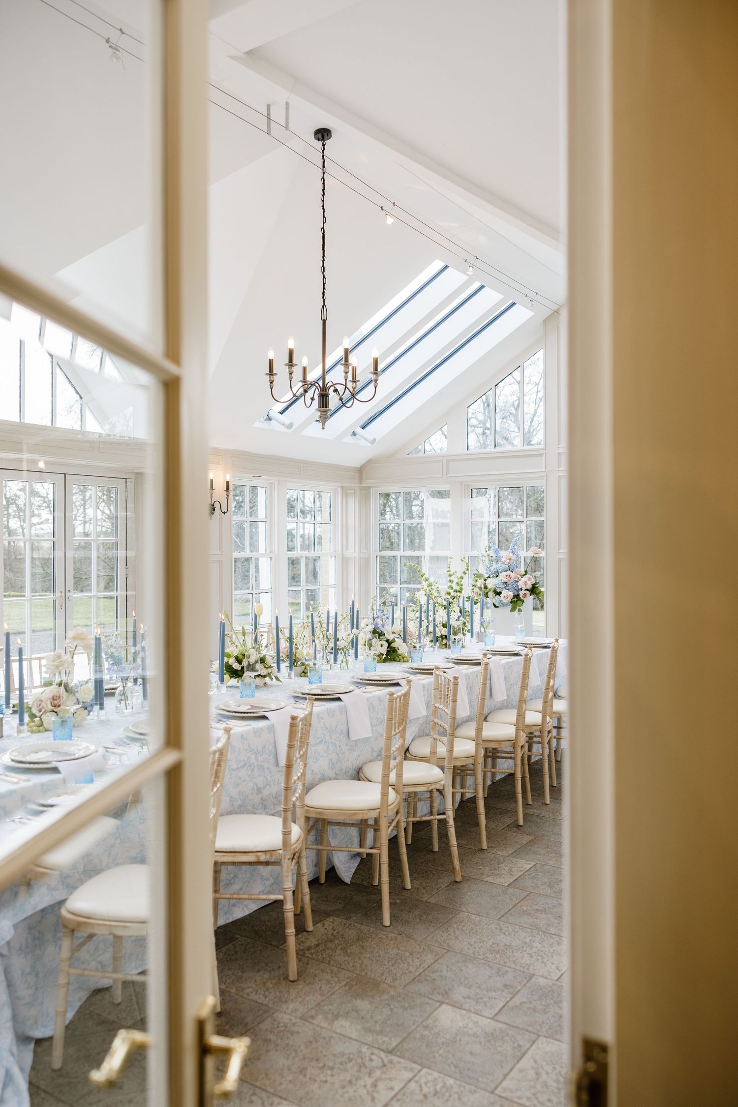 Elegant sunlit conservatory dining room with white chairs and tables arranged in a row beneath slanted glass ceiling panels.