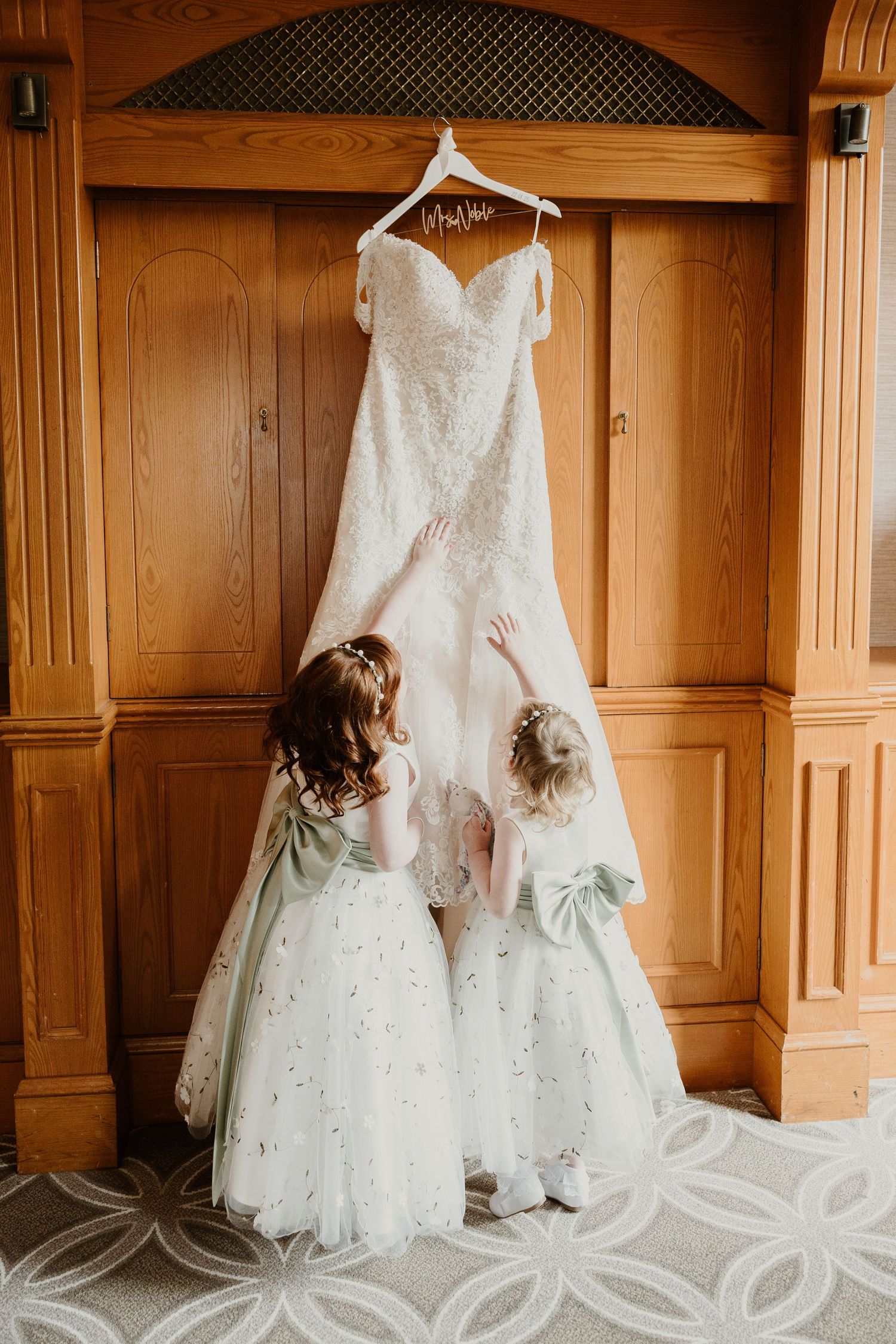Two young children in white dresses admire an elegant wedding gown hanging in a wooden room with paneled walls.