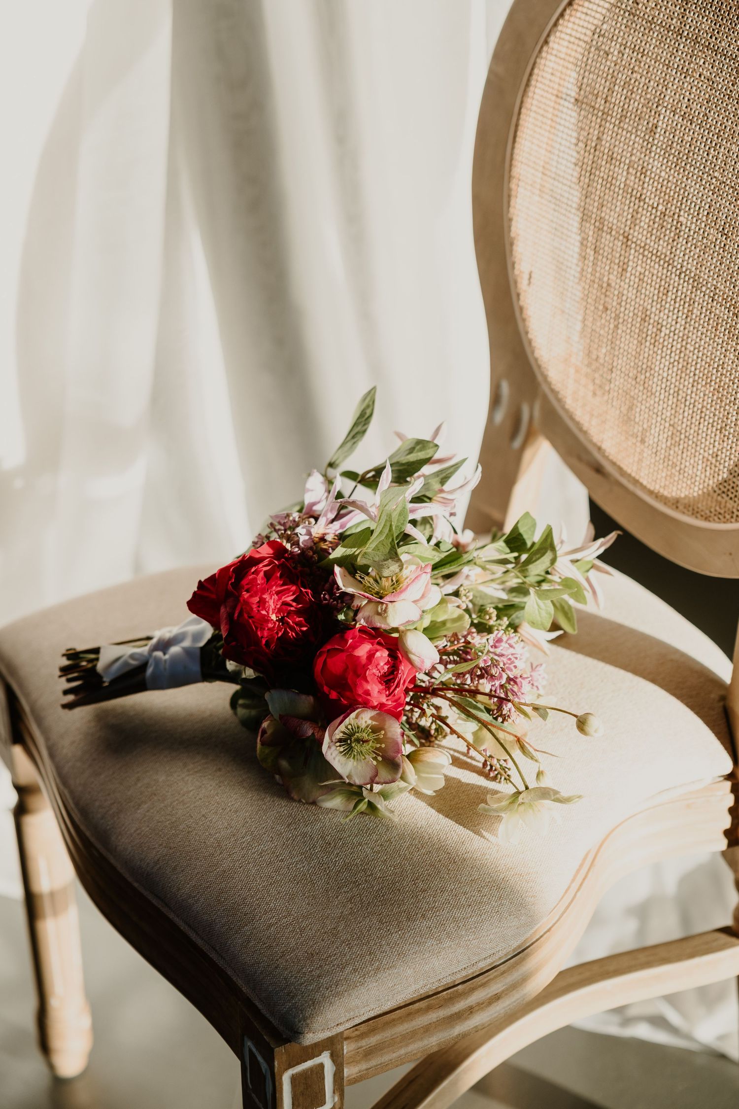 Red roses and floral arrangements rest delicately on vintage French cane chair against white backdrop.
