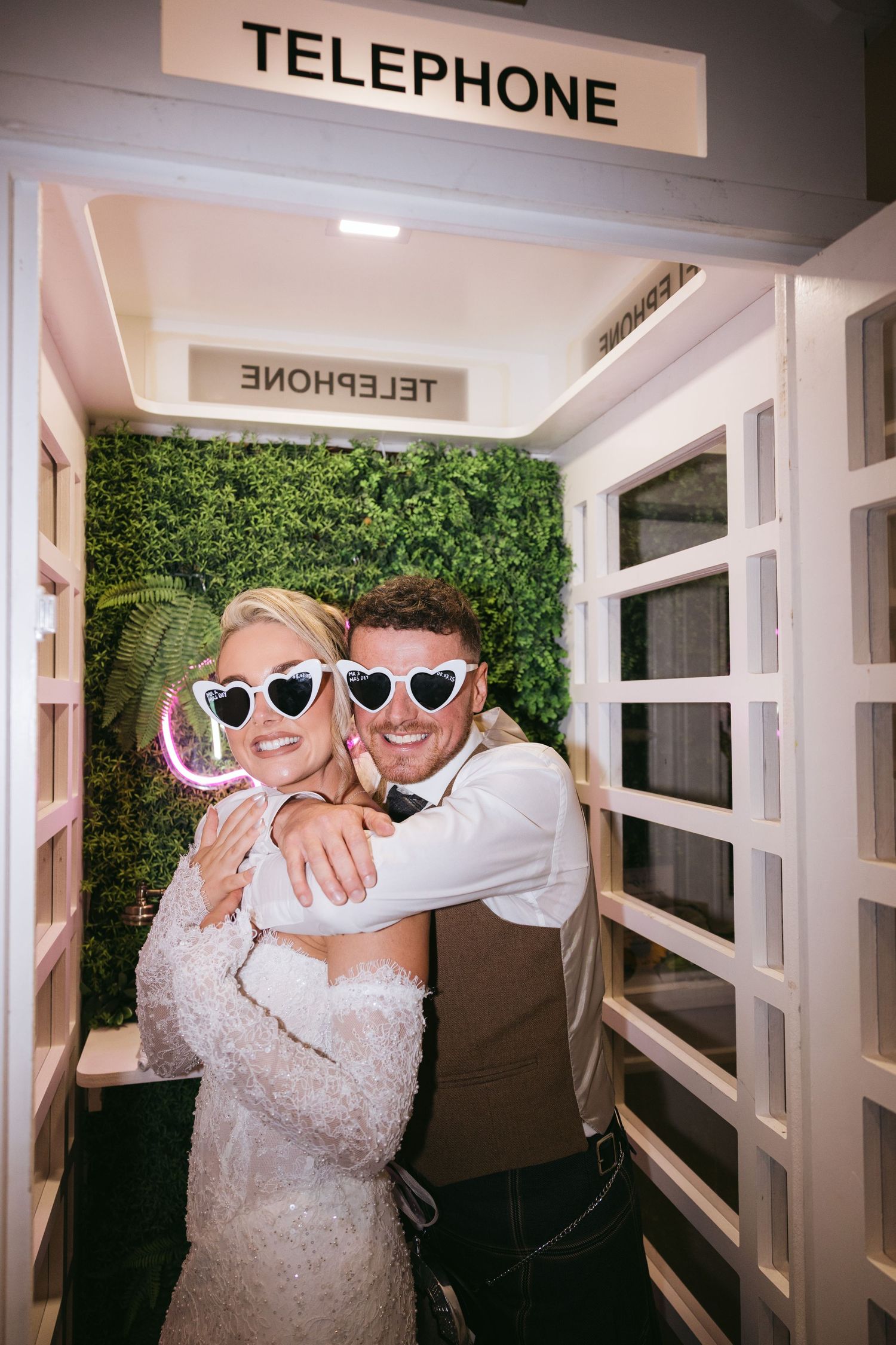 A couple wearing matching sunglasses pose together in a telephone booth with a green hedge wall backdrop.