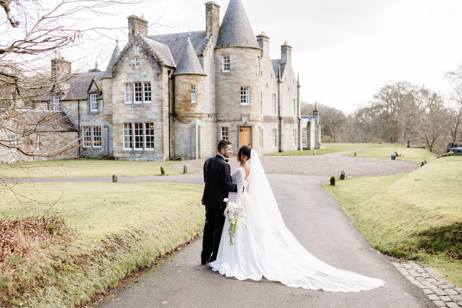 Elegant wedding couple poses in front of a historic stone mansion on a scenic countryside estate.