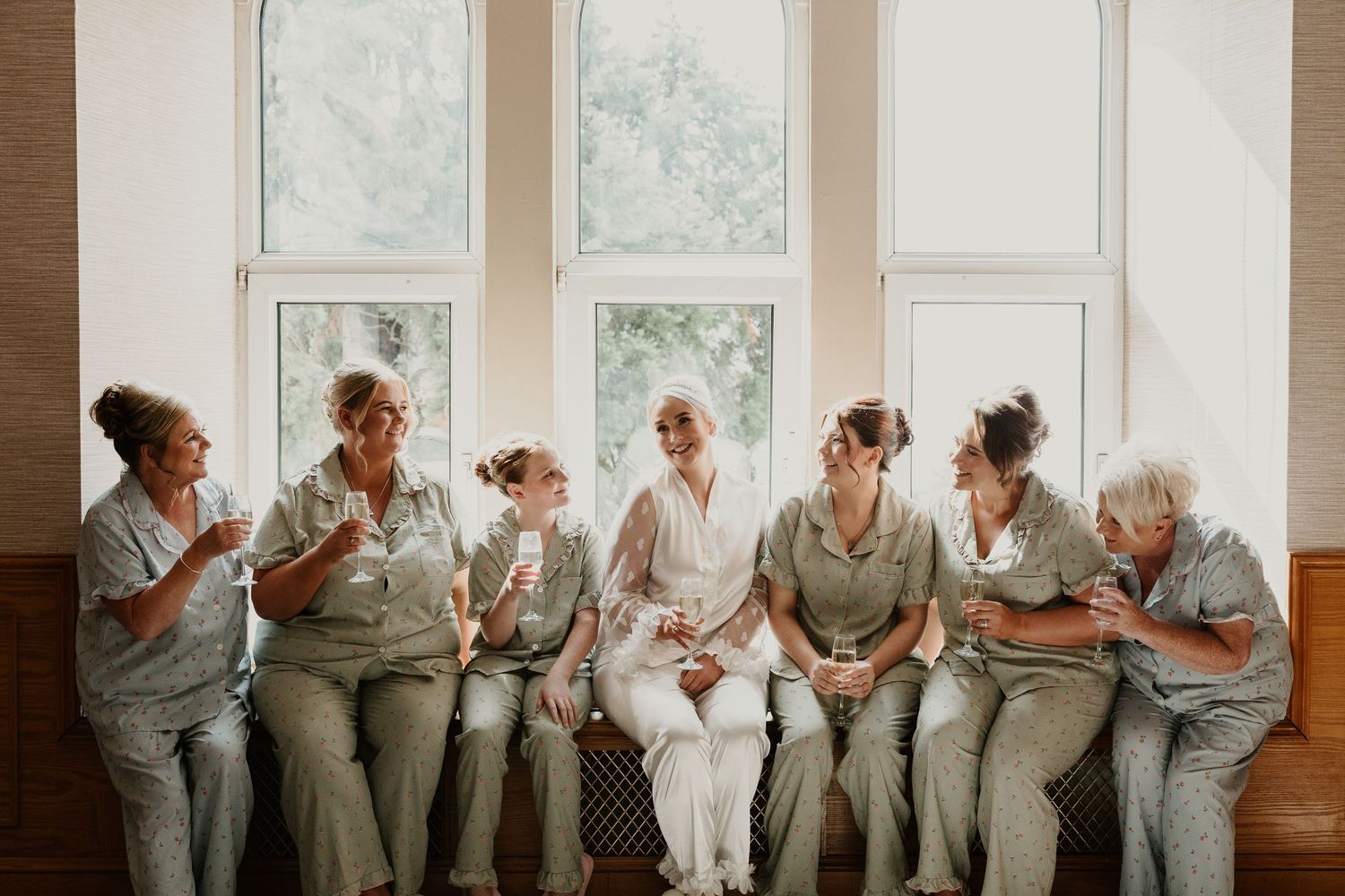 Bridal party in matching gray dresses sits on white window seats while checking their phones.