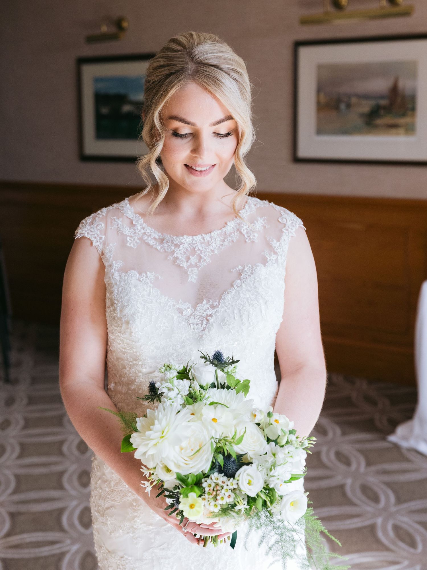A radiant bride in a delicate lace wedding gown holds an elegant white and green bouquet while looking down with joy.