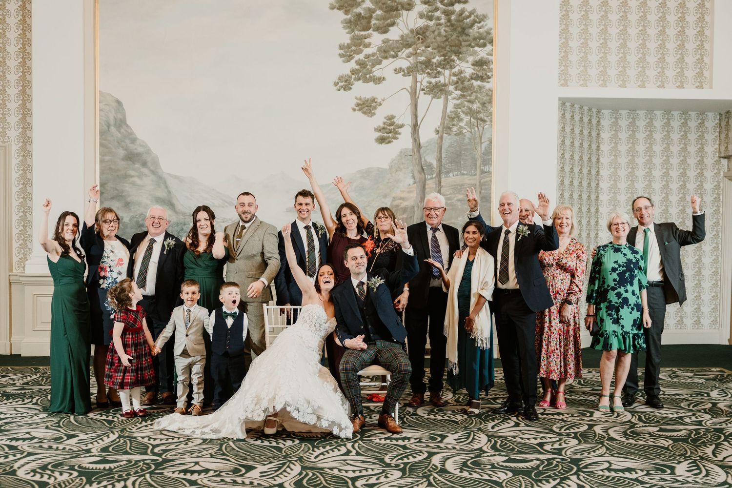 Large wedding party poses together outdoors in a mix of burgundy and green formal attire while celebrating.