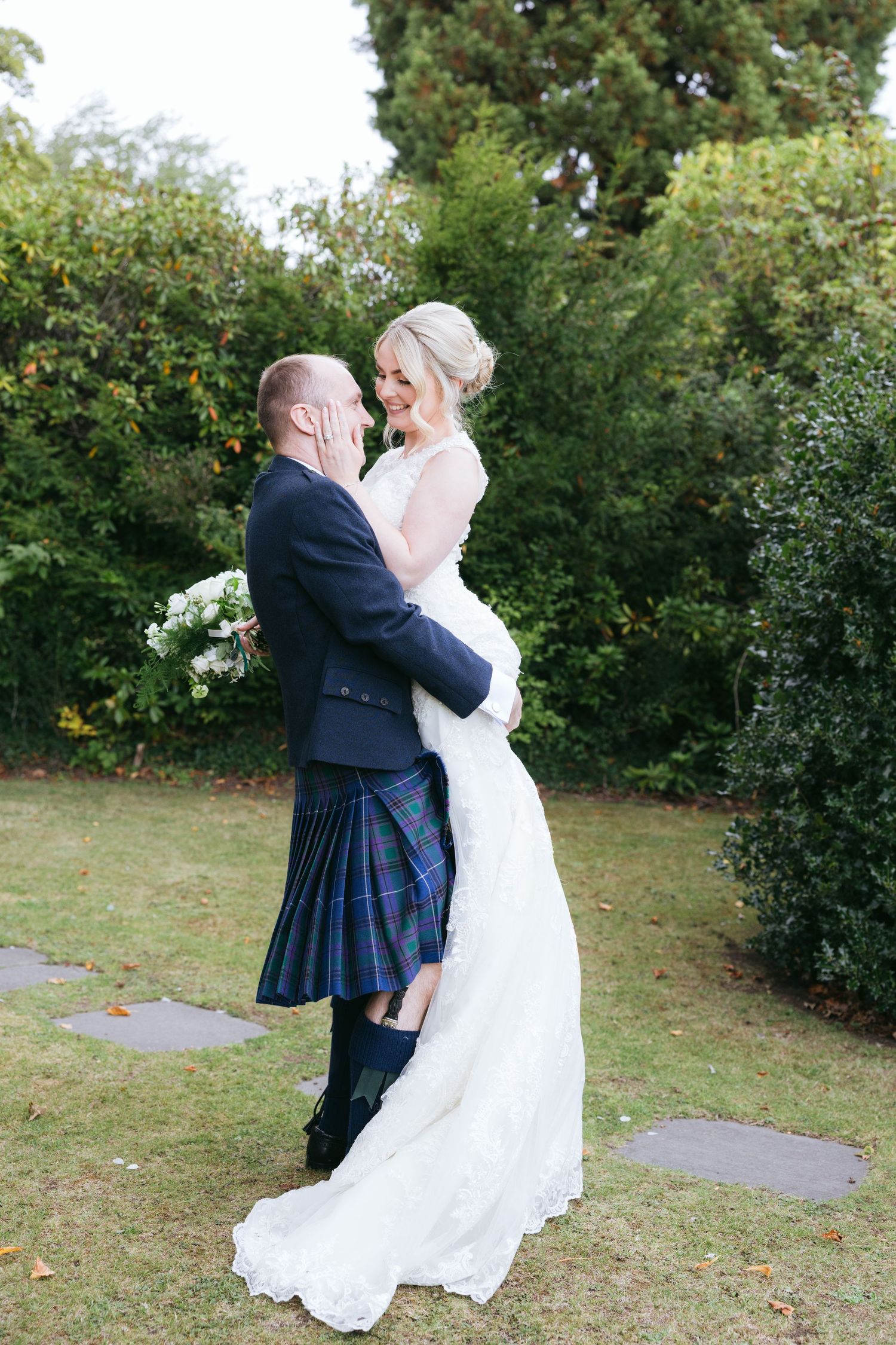 A romantic wedding couple in navy kilt and white dress embrace outdoors on grassy lawn near garden foliage.