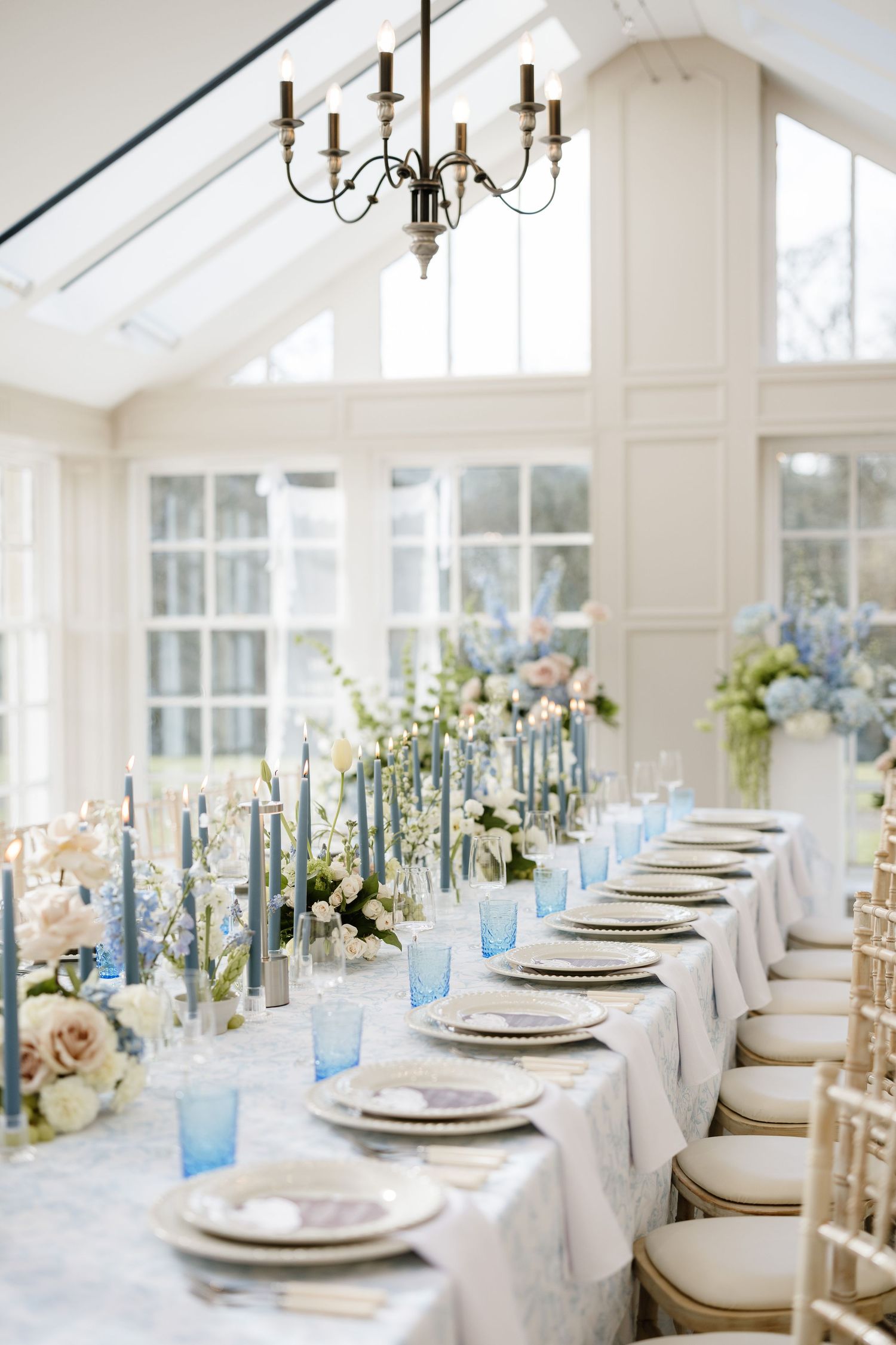 Elegant wedding table setting with blue glassware, white linens, and brass candelabras in a bright sunlit venue.