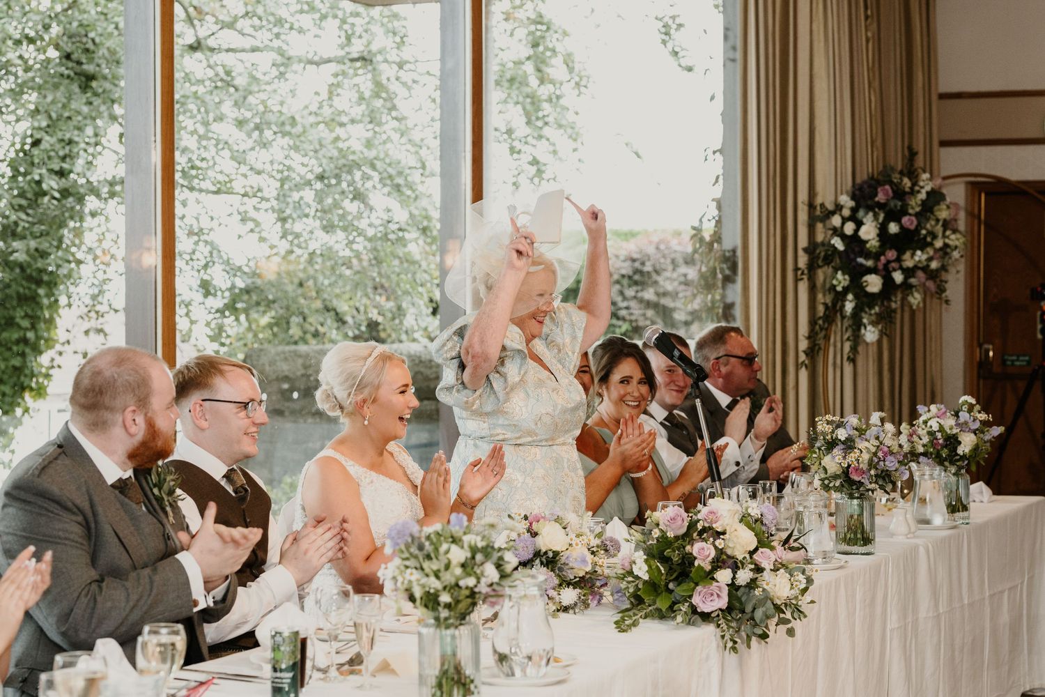 Wedding guests raise their glasses in a festive toast during an elegant reception celebration with floral centerpieces.
