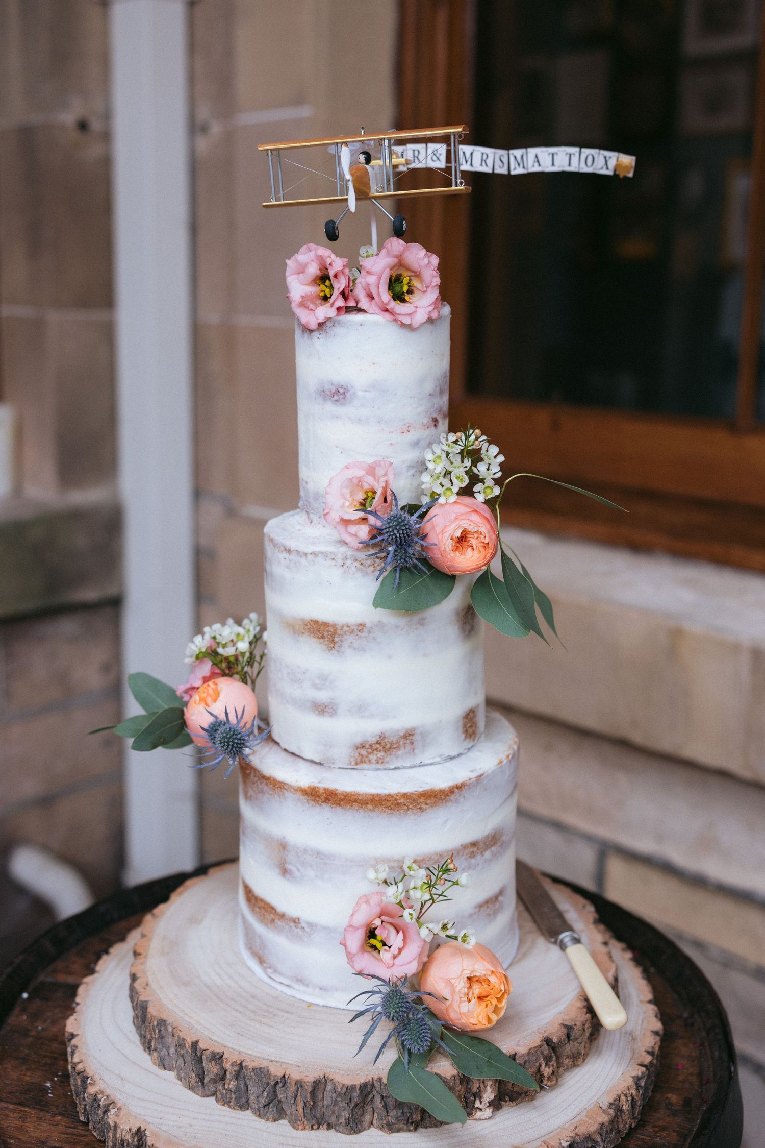 A rustic semi-naked wedding cake decorated with fresh flowers and greenery, displayed on a wooden slice cake stand.