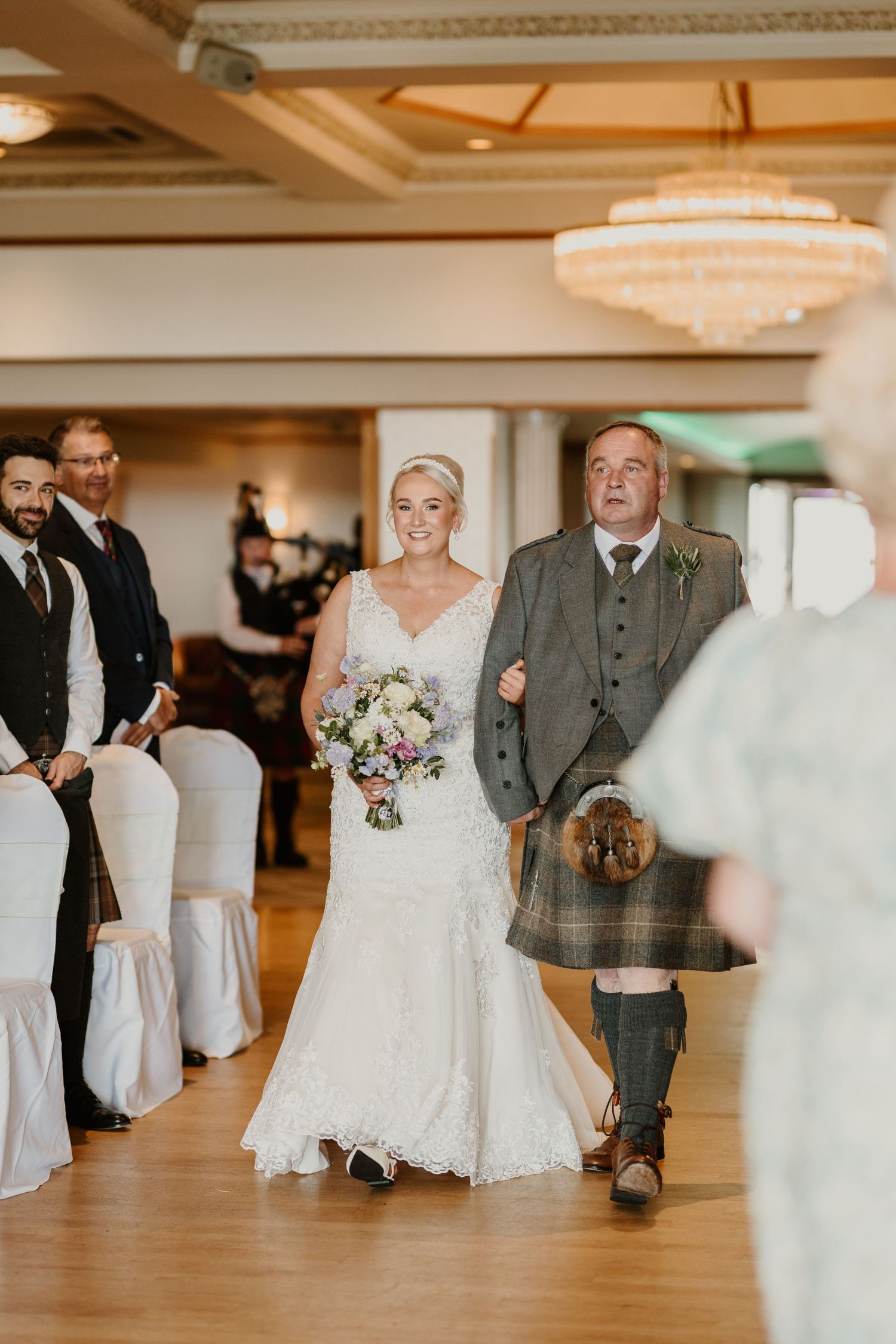A bride and her escort in traditional Scottish kilt walk down the aisle at an elegant indoor wedding ceremony.