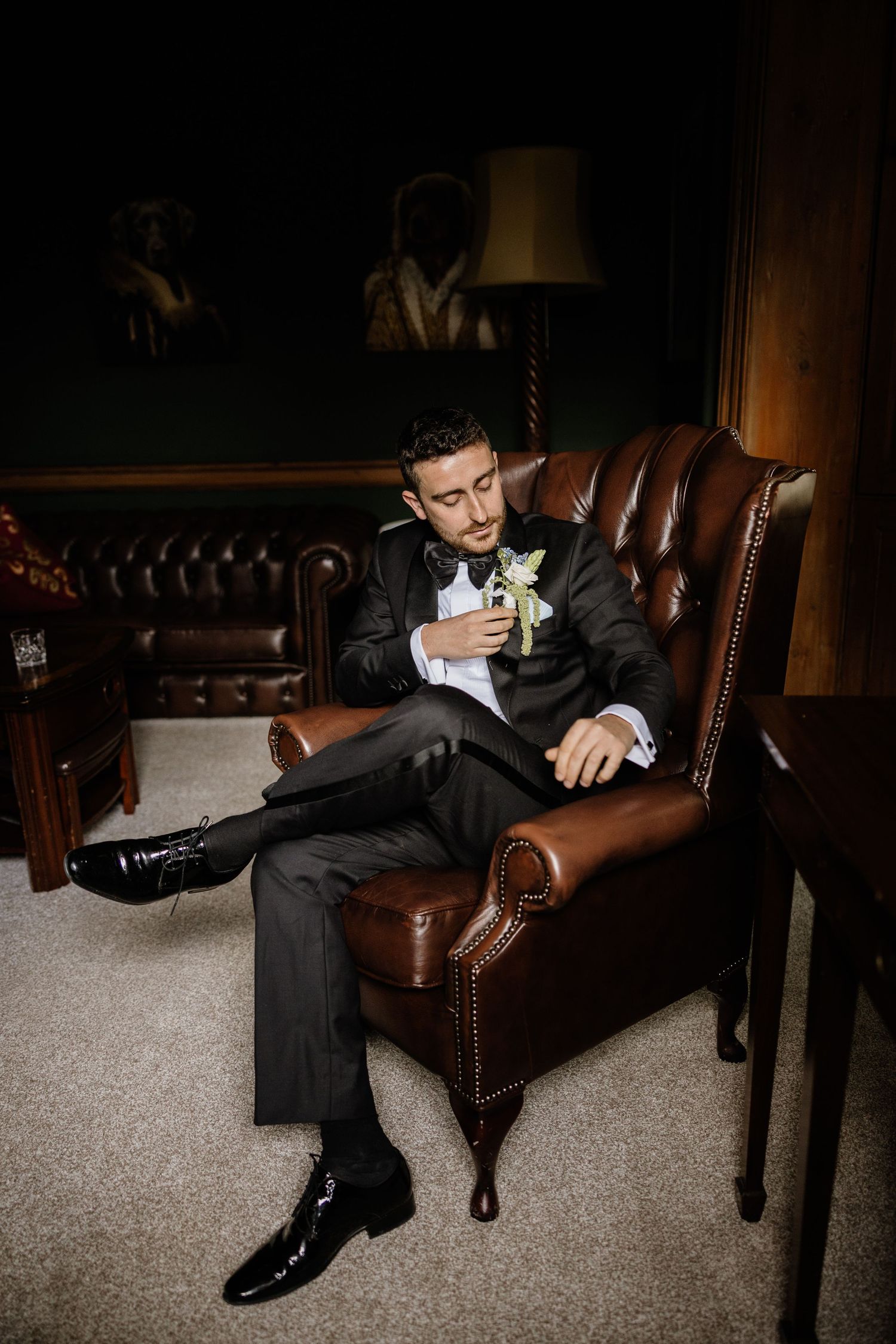 Person in formal black suit lounges on brown leather armchair in dark moody setting.