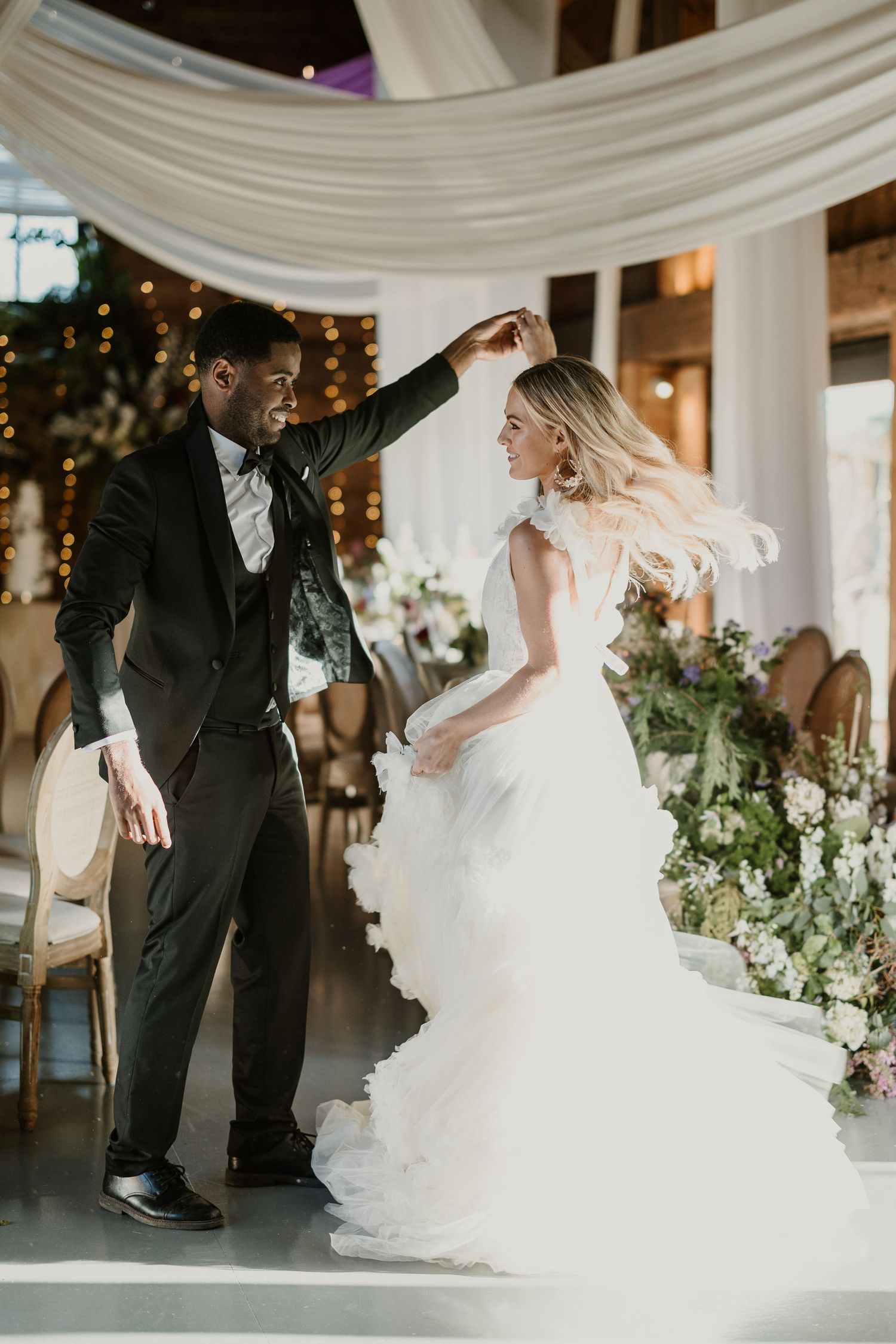 A couple shares a romantic dance moment under white draped fabric and twinkling lights.