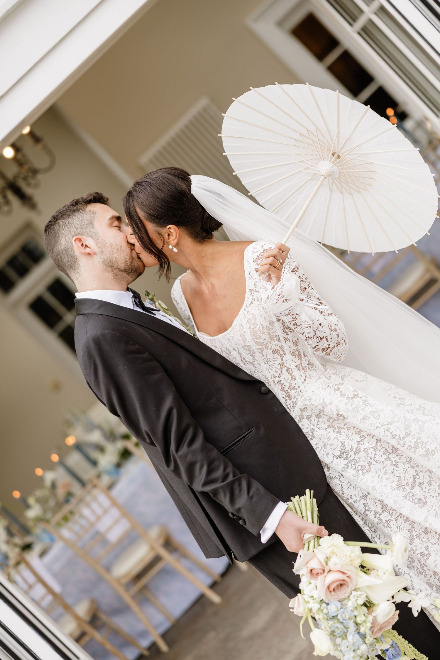 A romantic wedding couple shares an intimate moment under a white parasol with a bouquet of pink roses.