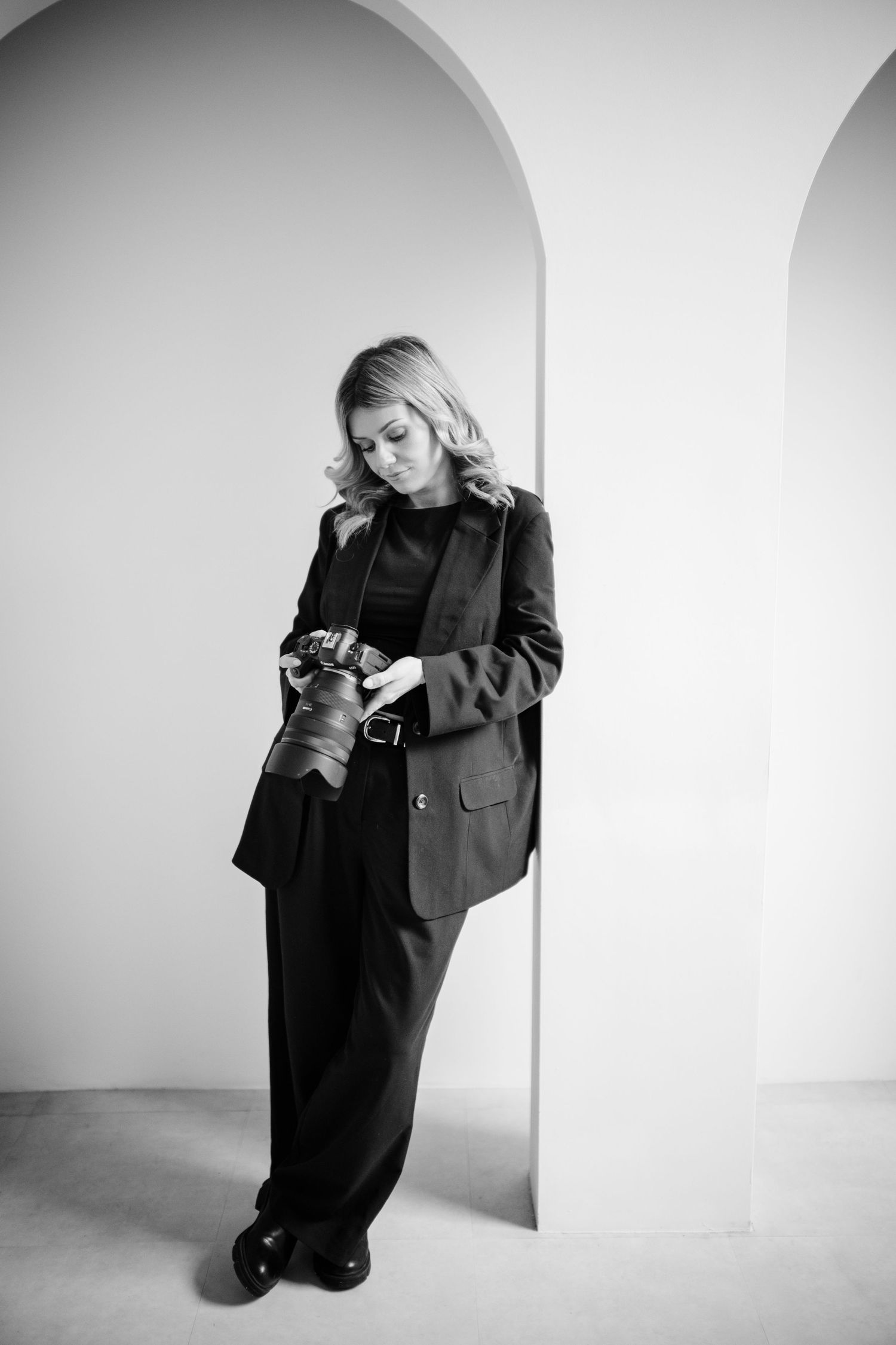 Black and white portrait of person in business attire looking at phone while standing against white arched wall.