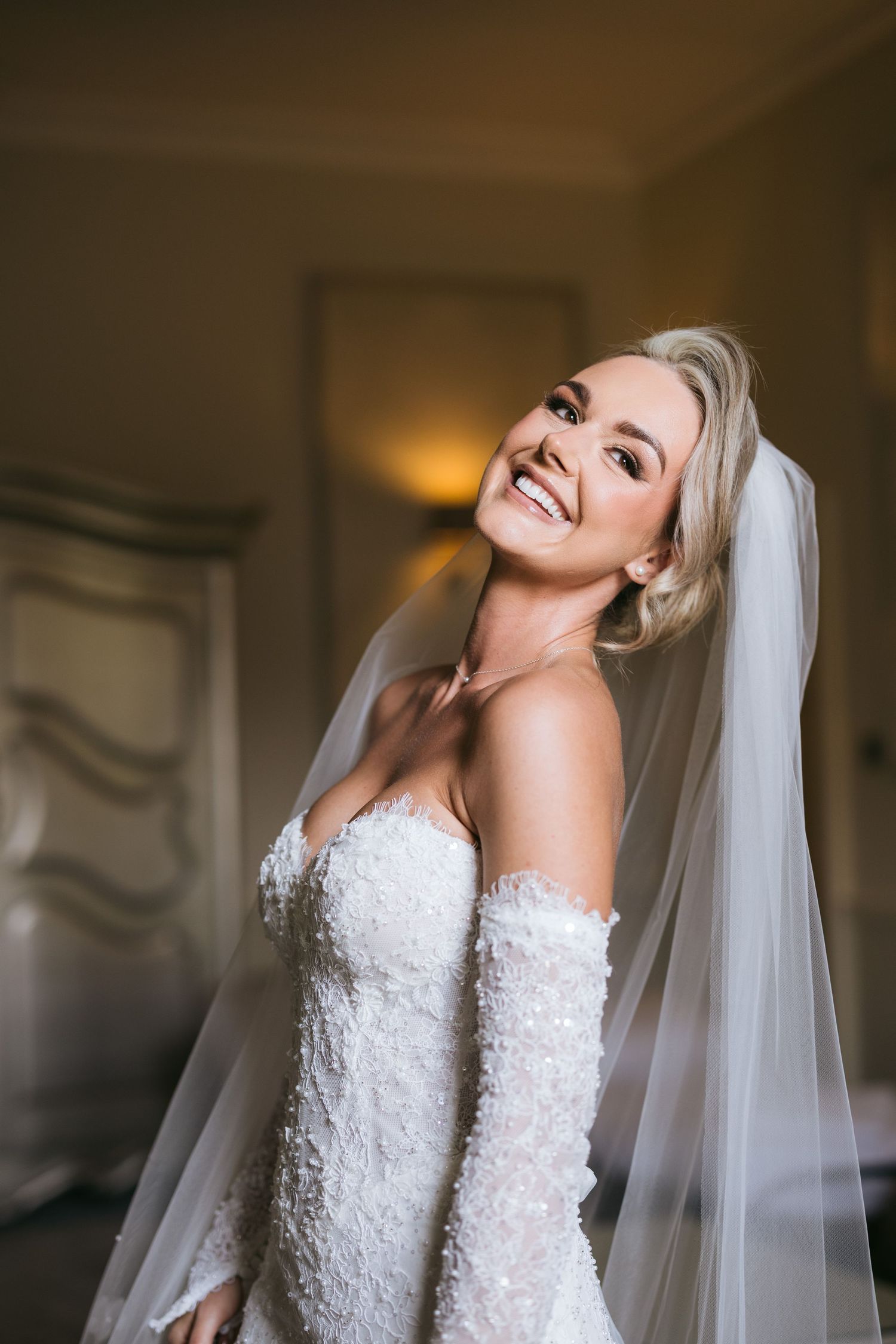 A bride in a white lace off-shoulder wedding dress laughs joyfully while wearing a flowing veil.