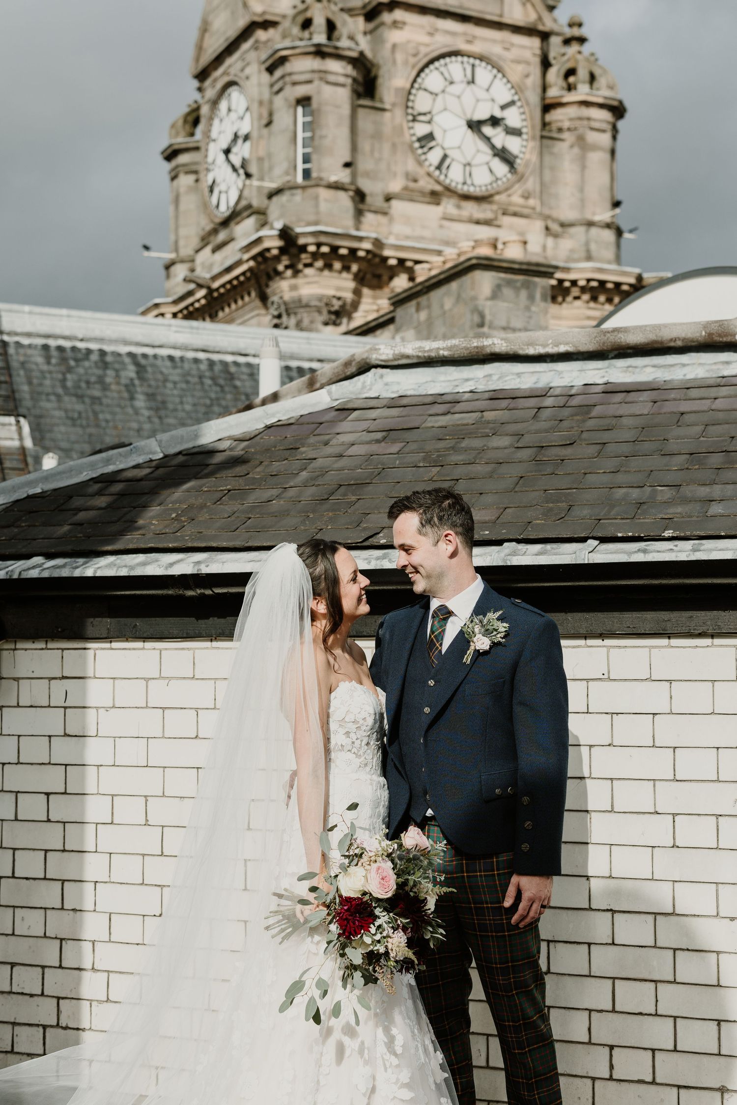 Newlyweds share a romantic moment on rooftop with historic clock tower and traditional Scottish architecture in background.