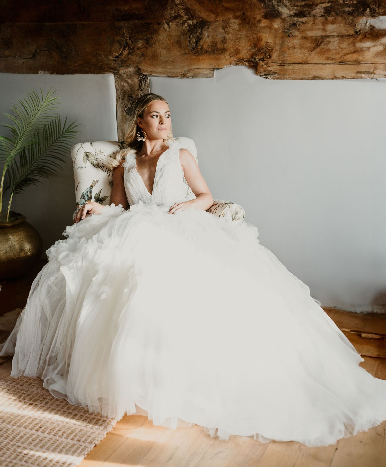 A bride in a voluminous white tulle wedding gown lounges gracefully on a rustic wooden bench in natural lighting.