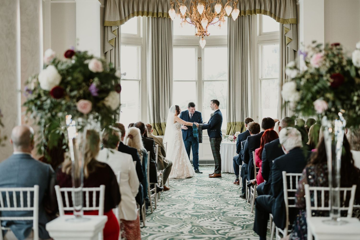 An elegant ceremony venue with high ceilings, chandeliers and floral arrangements showcasing guests seated in rows.