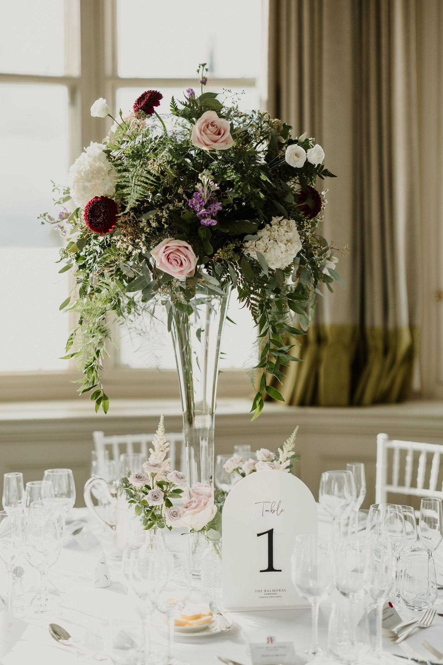 Elegant wedding centerpiece with pink and burgundy flowers in tall glass vase on white tablecloth.