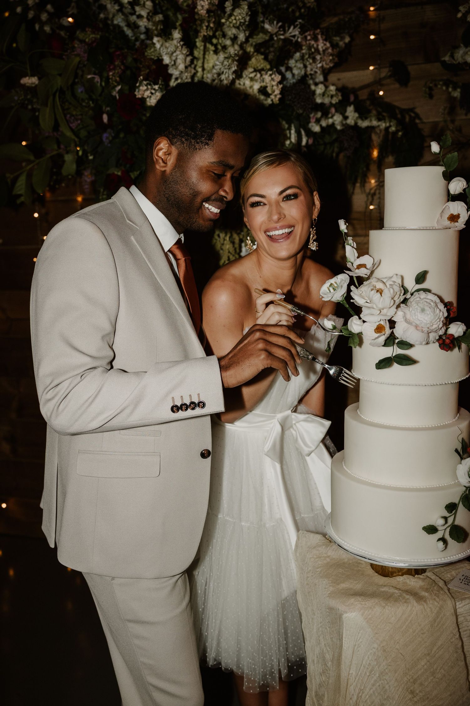 A joyful wedding couple cuts their elegant white tiered cake decorated with greenery at their reception.