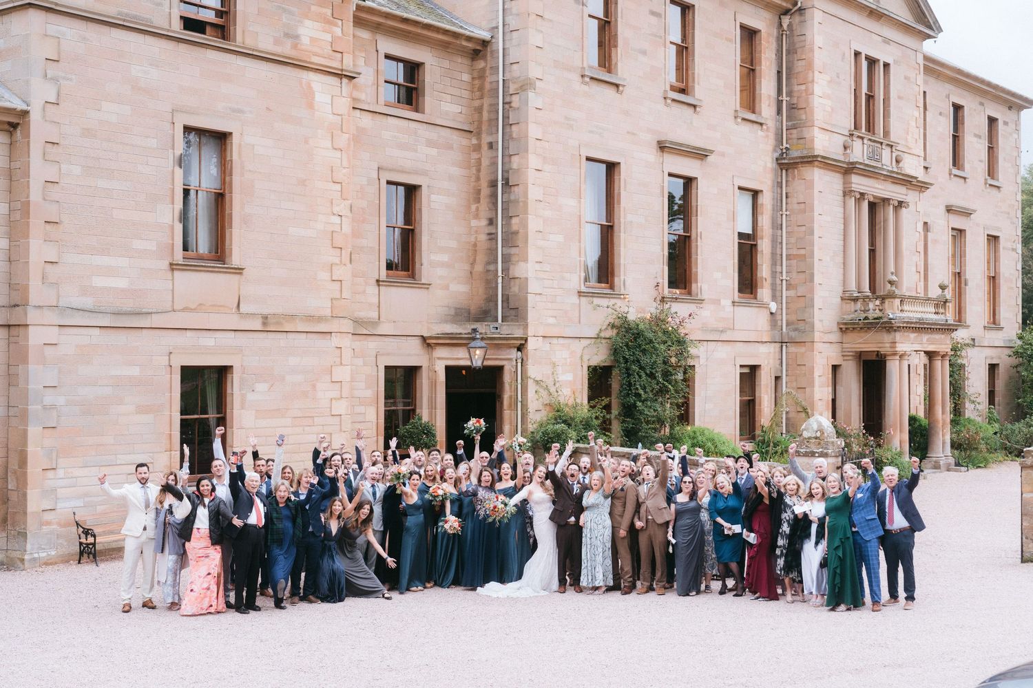 Large wedding party group photo gathered outside a grand stone building.