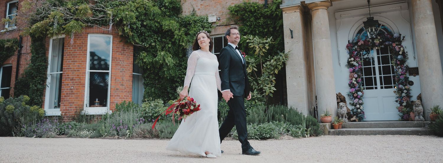 A couple in wedding attire walks together outside a historic brick building with arched windows.