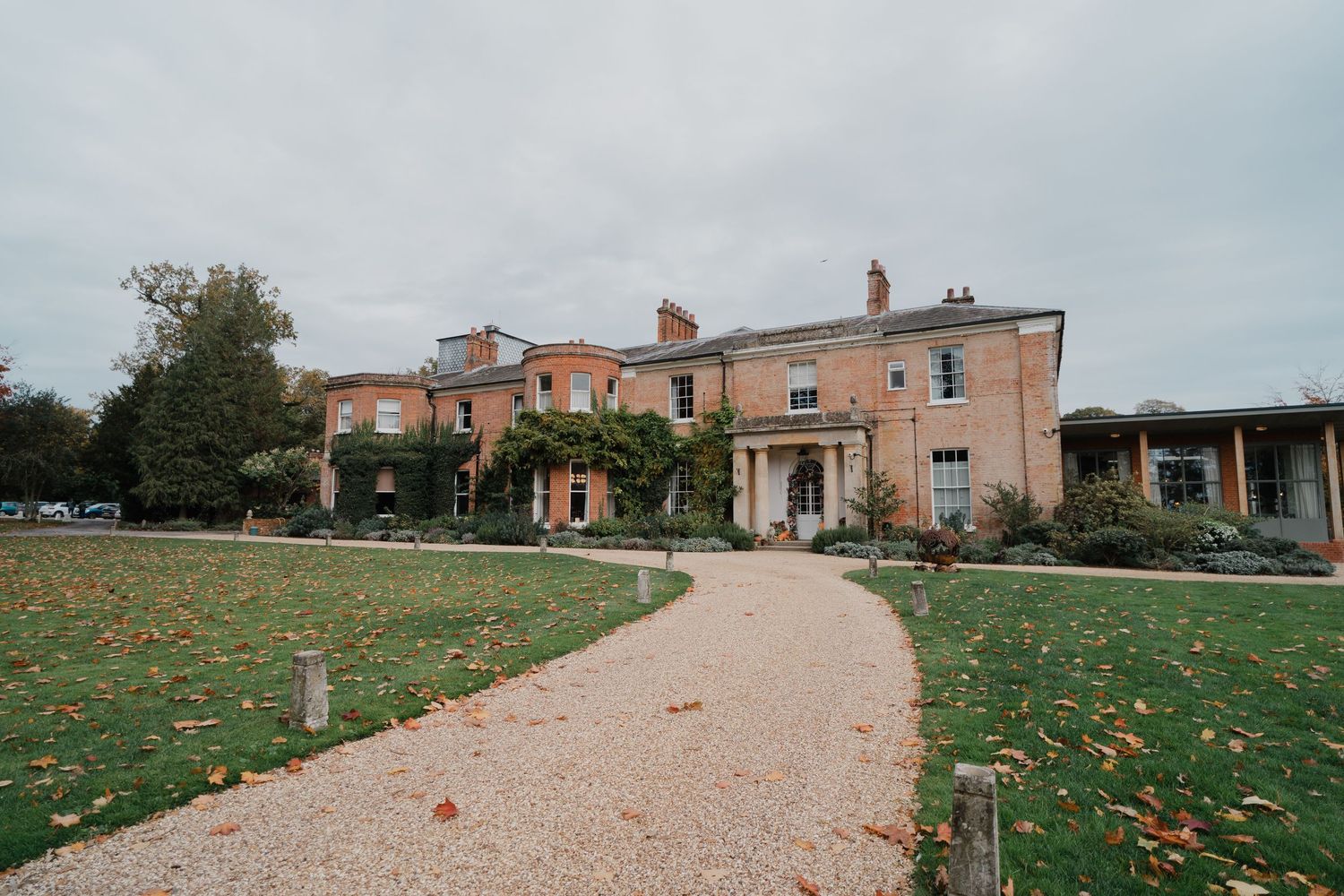 A gravel pathway leads to a pink Georgian manor house with autumn leaves scattered on the surrounding lawn.