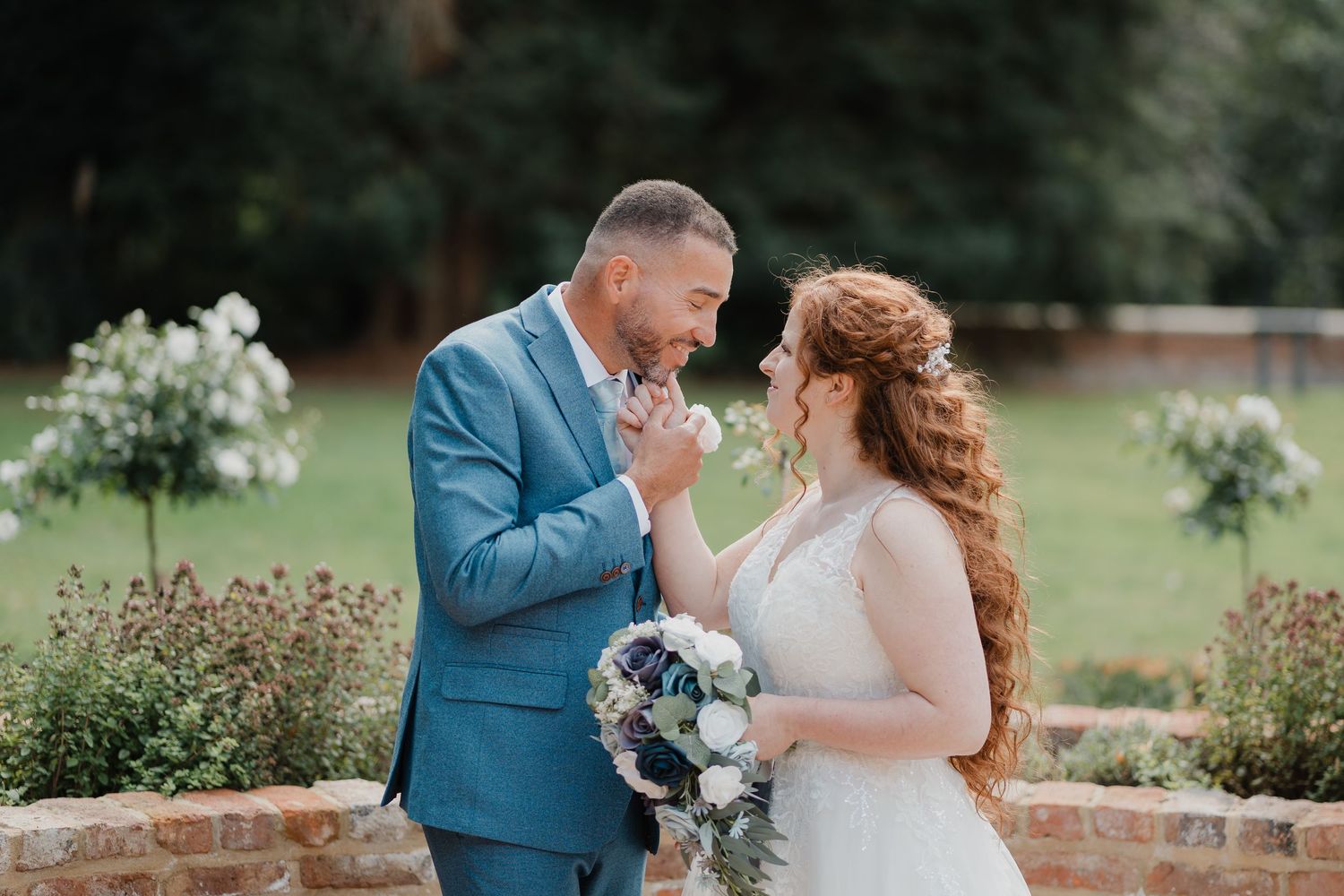 A romantic couple in formal attire shares tender moments during their outdoor wedding photoshoot in a garden setting.