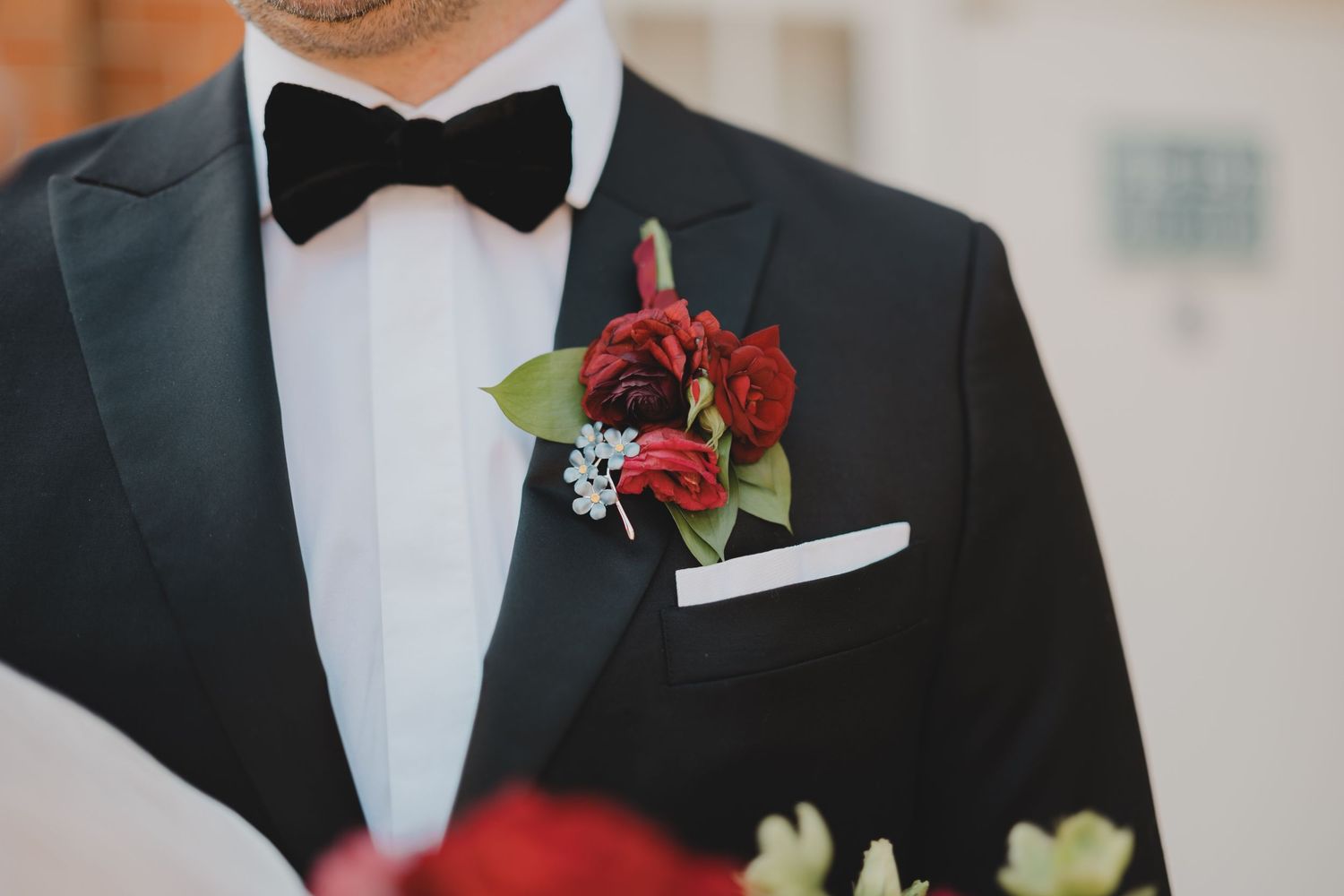 A groom in a black tuxedo with a red rose boutonniere stands at a formal wedding event.