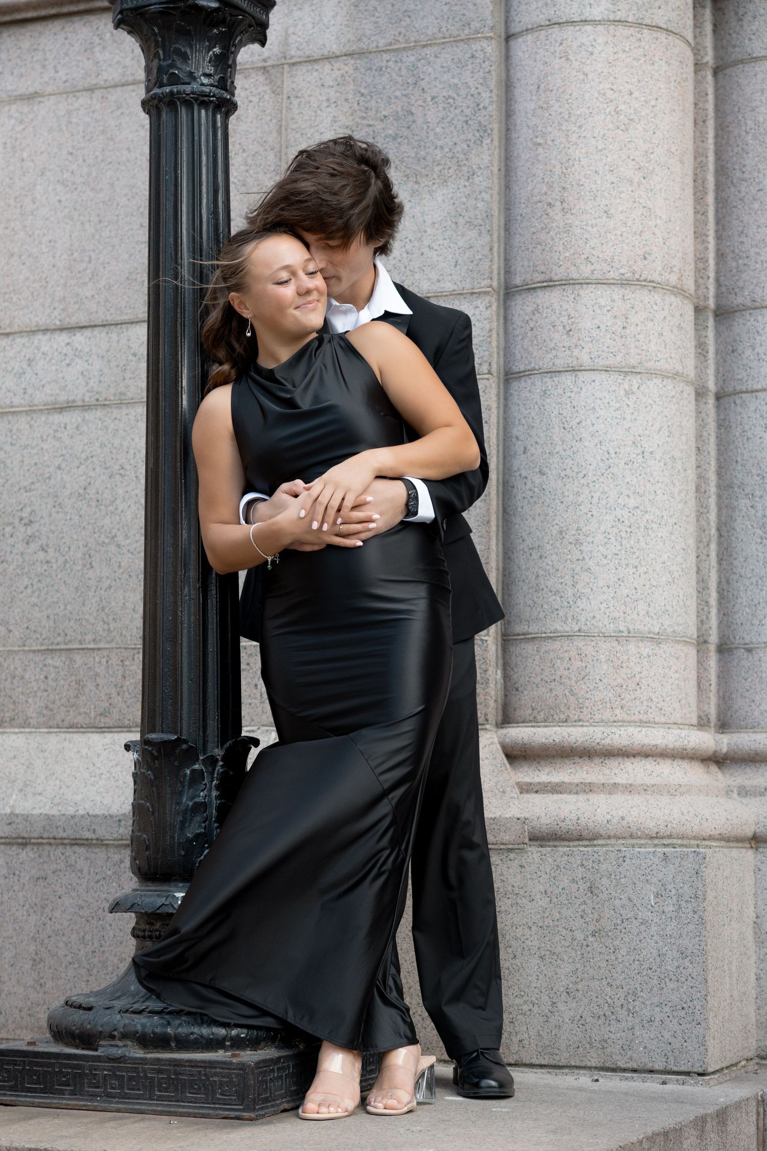 A romantic couple in formal attire embraces by a stone pillar, with the woman wearing an elegant black evening gown.